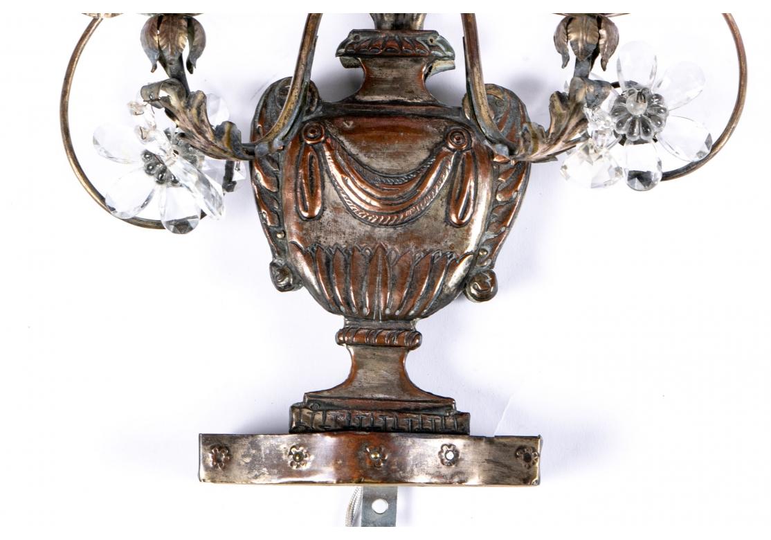 A pair of classically themed Sconces with Footed draped urn forms with tall scrolled leafy vines with 5 crystal flowers. The sheet metal urns attached to wood. The twin scrolled lights with crystal pendants. 
Measures: Height. 20