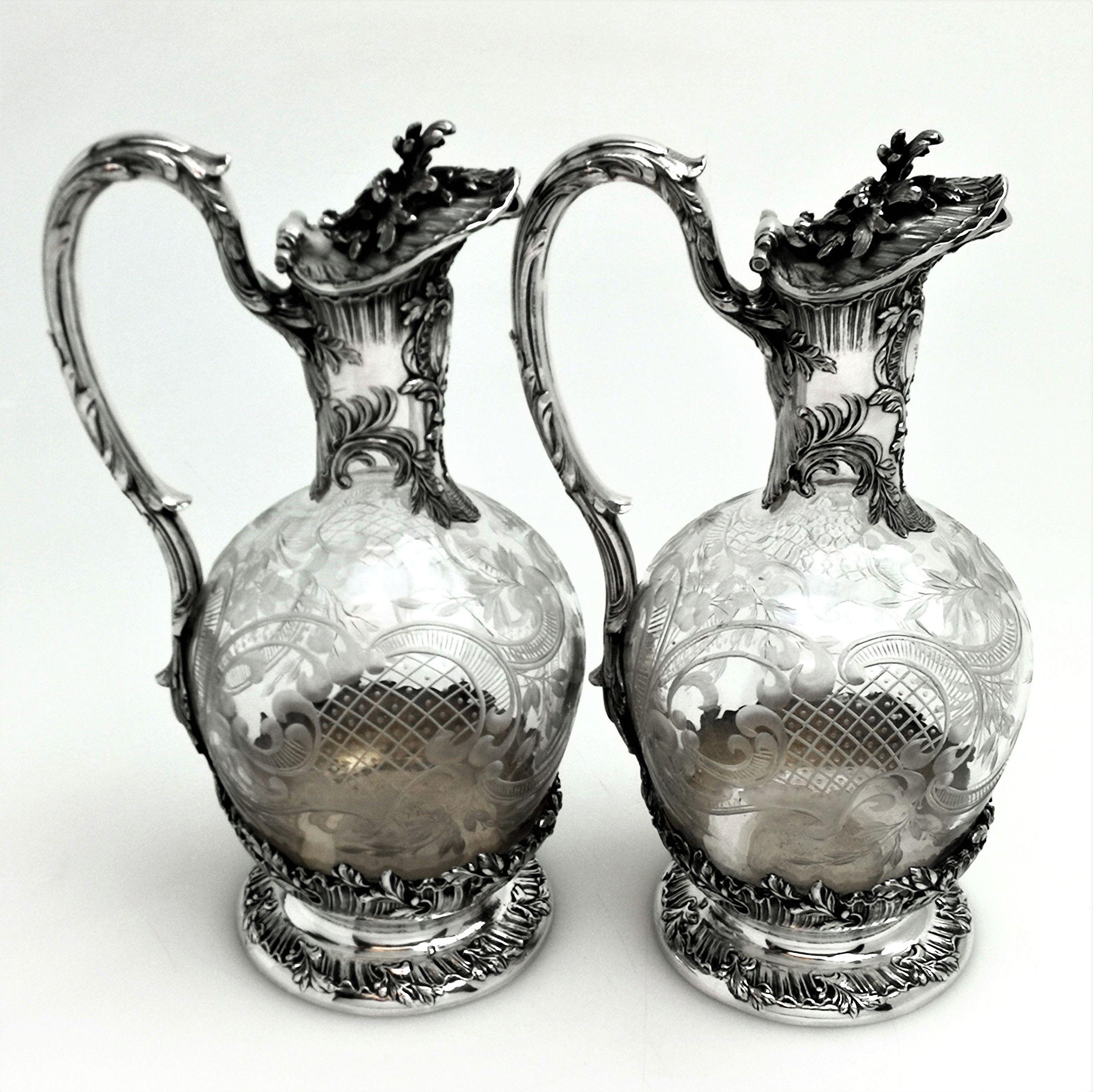 A beautiful pair of antique French solid silver and glass claret jugs. These wine jugs feature ornate chased patterning including a shaped cartouche with an engraved monogram below the spout. The Jugs each have a foliate topped handles that reach