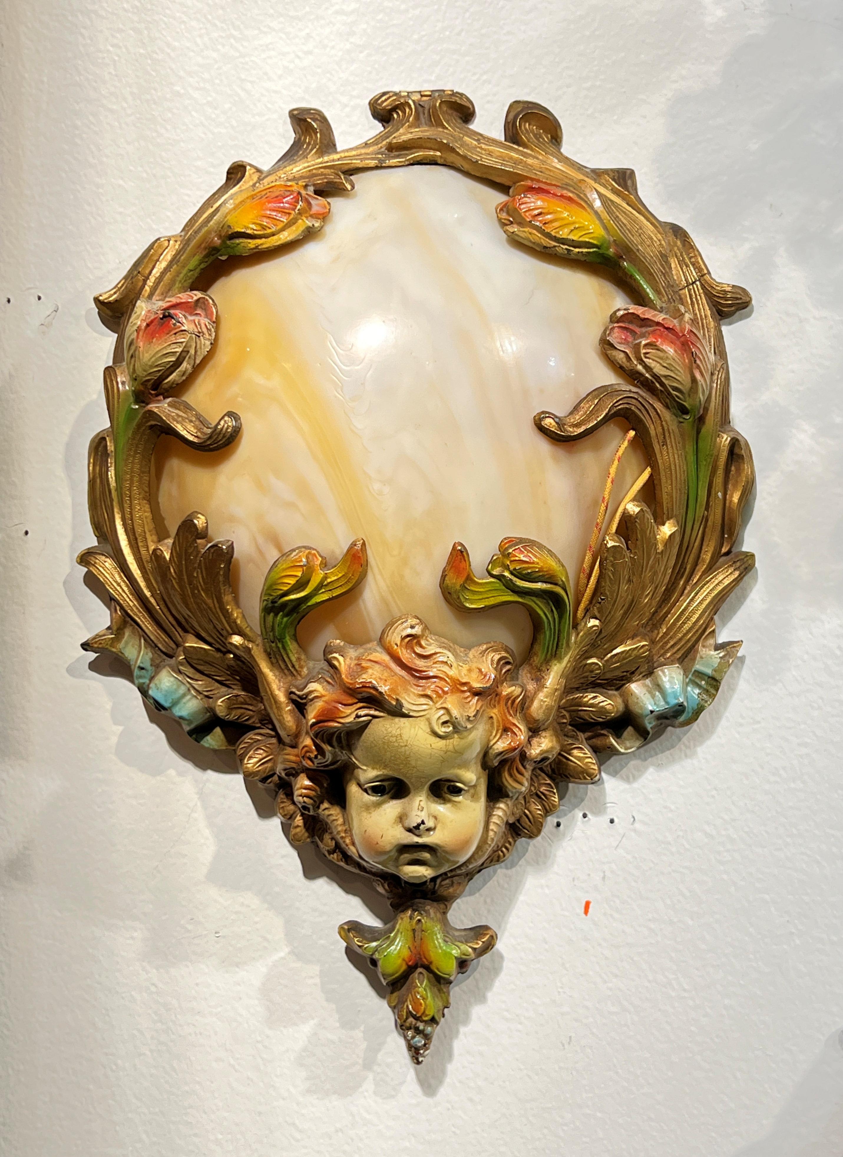 Our unique pair of Art Nouveau period antique sconces with dome shape are crafted from blown molded variegated white, cream and gold colored glass with cold-painted bronze frame depicting blooming flowers with neoclassical cherub mask at the base. 