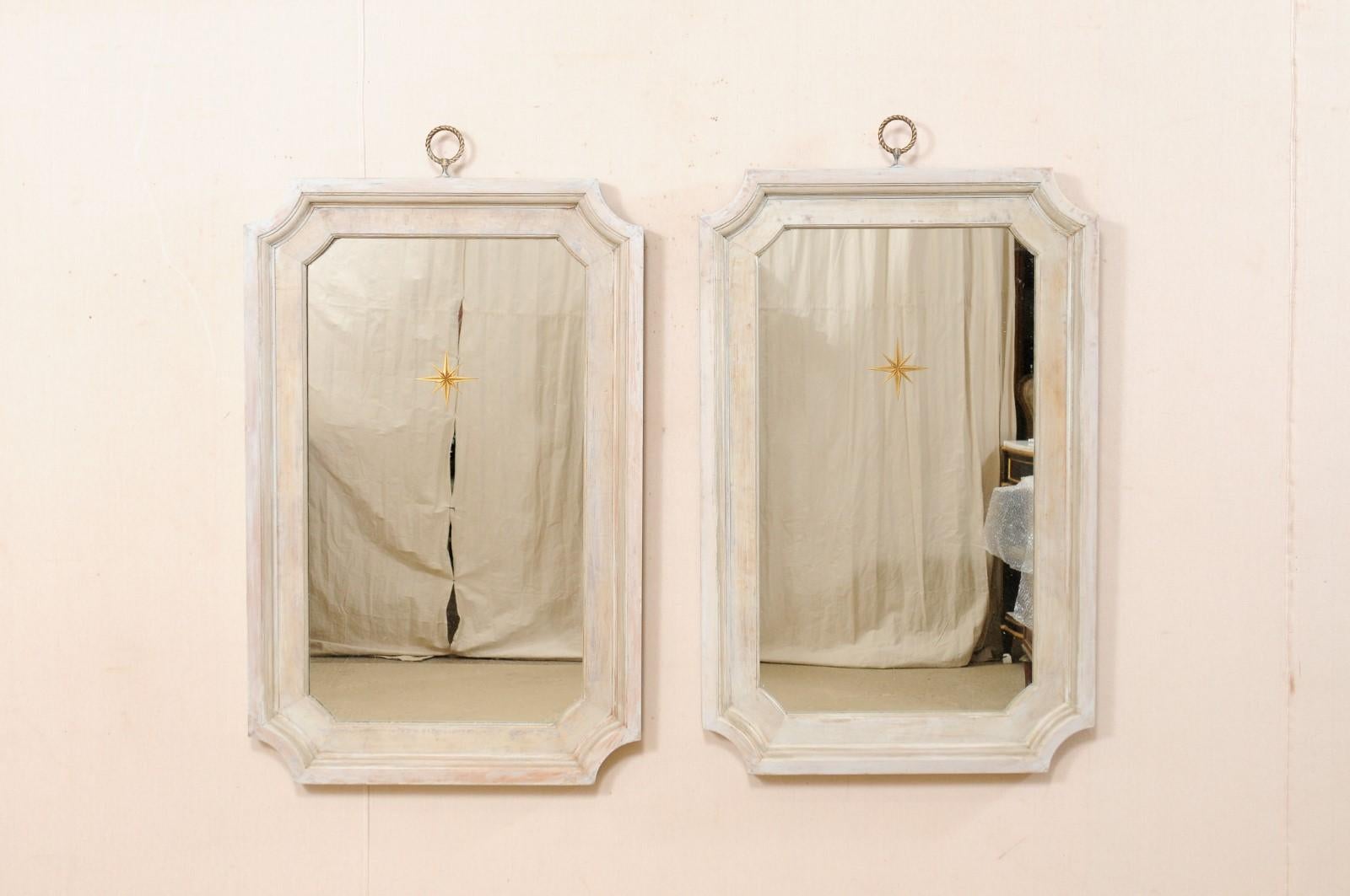 A pair of Artisan-made mirrors with gold églomisé sunbursts, within carved and painted wood frames. This pair of mostly rectangular-shaped mirrors have been artisan crafted with a clear mirrored center, which is minimally adorn with a single gold