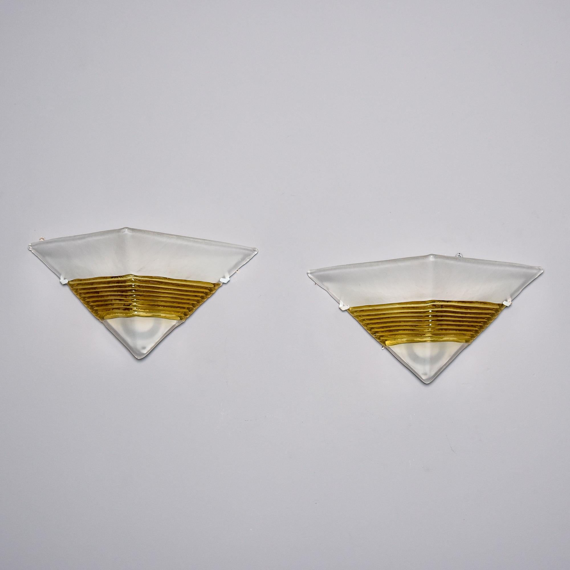 Vers les années 1990, paire d'appliques en verre de Murano par AV Mazzega. Chaque abat-jour triangulaire en verre blanc épais et presque opaque est rehaussé d'une bande dorée striée. Les abat-jours sont maintenus en place au ras du mur et recouvrent