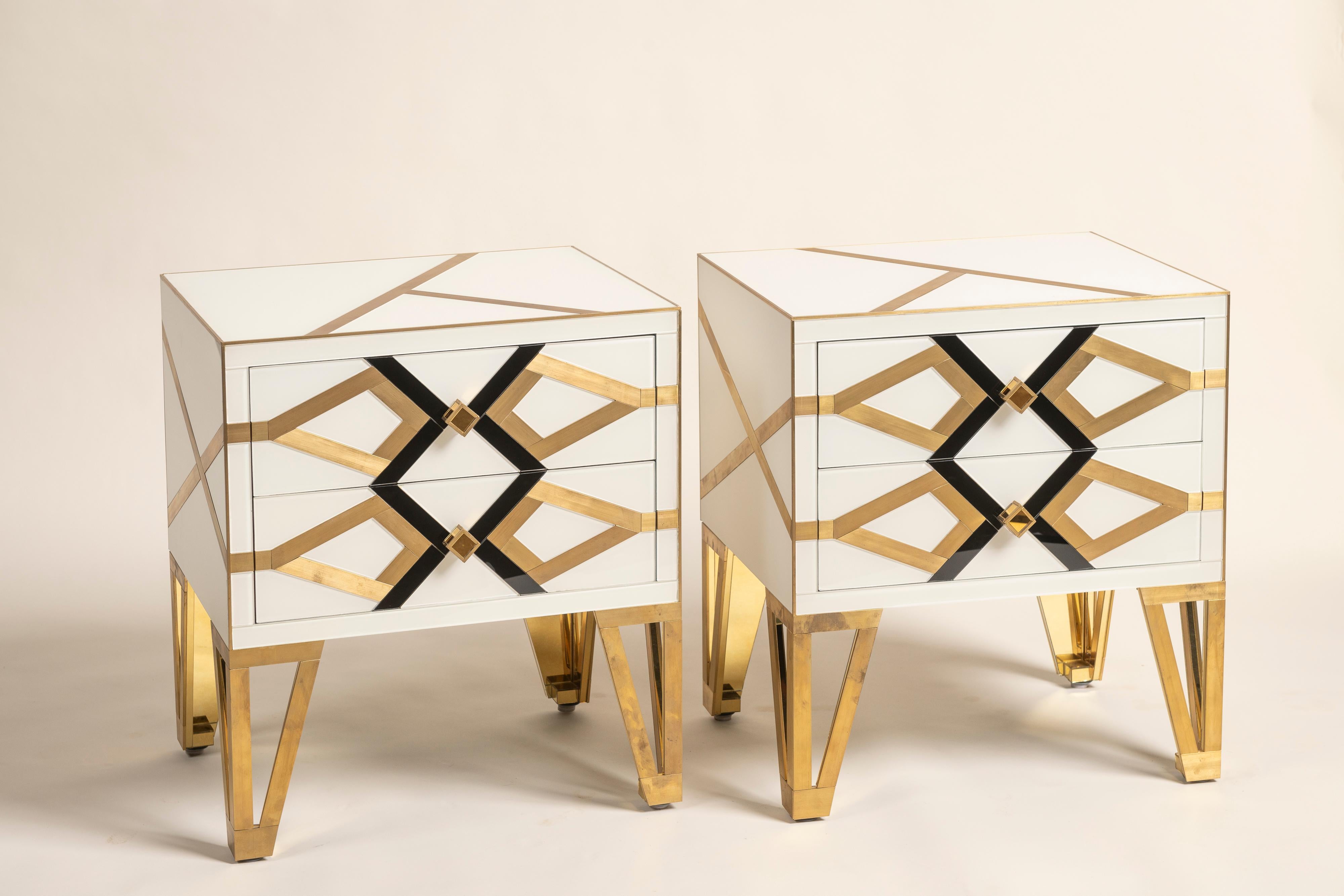 A striking pair of Italian modern two-drawer nightstands/side tables with an Art Deco flair — unique, handmade pieces by Cosulich Interiors & Antiques. The ivory white frame features a bold black glass geometric pattern, with brass gold inserts and