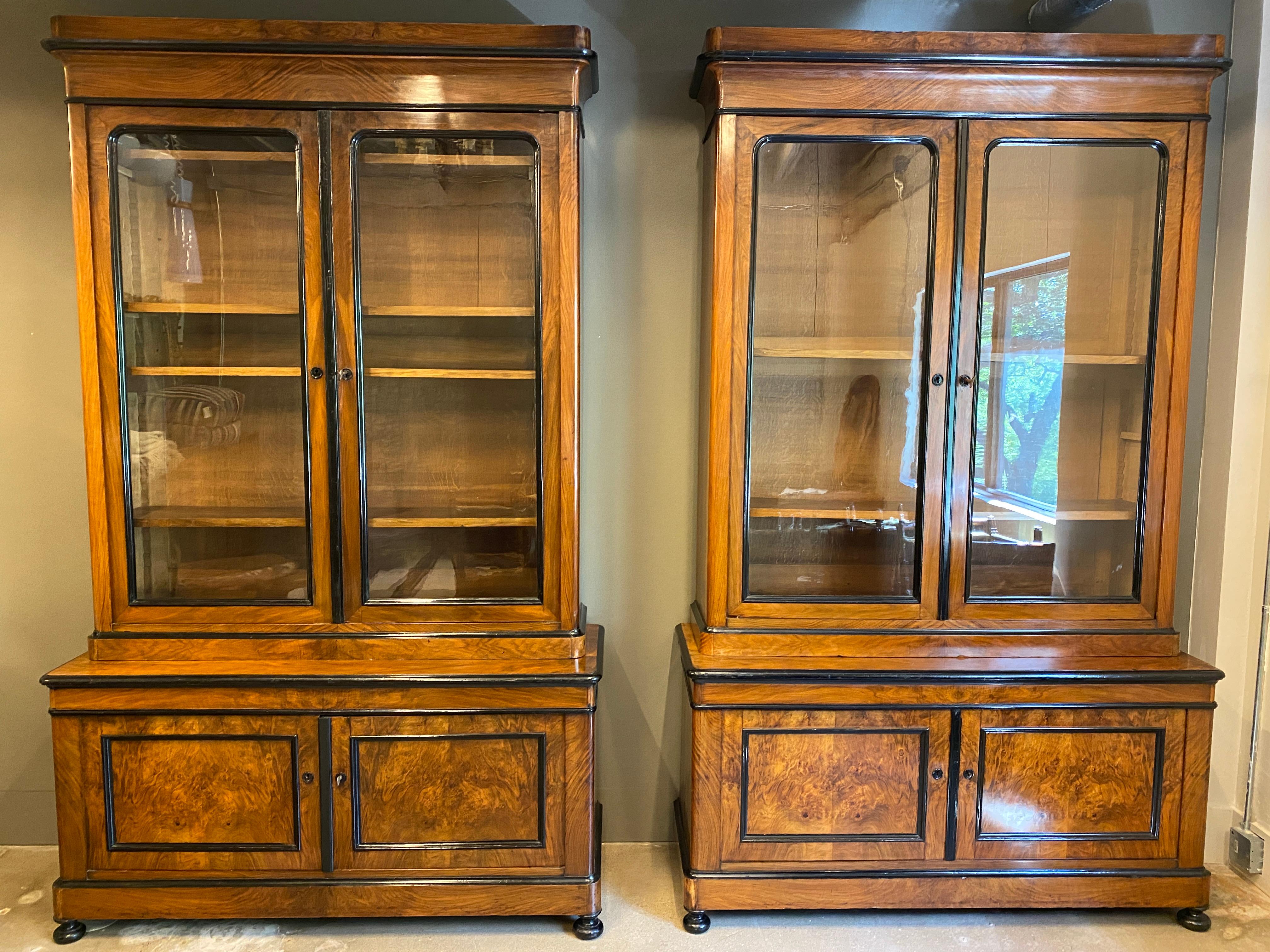 Hard to find pair of large scale Austrian Biedermeier step-back cabinets of burled walnut or fruitwood with black ebonized accents. Top section has double glass doors and four adjustable oak interior shelves. Base has panel doors and single interior