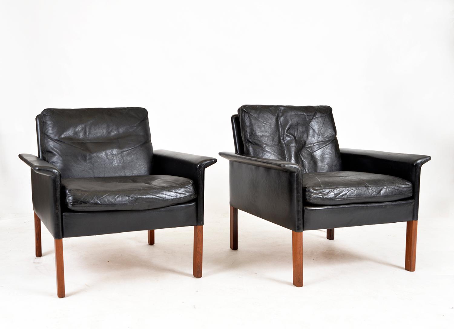 This handsome pair of 1960s black leather lounge chairs sat on square hardwood feet are part of the CS500 series designed by Hans Olsen in 1962 for Christian Sørensen & Co (C.S Furniture). 
These mid-century chairs have been well loved over the