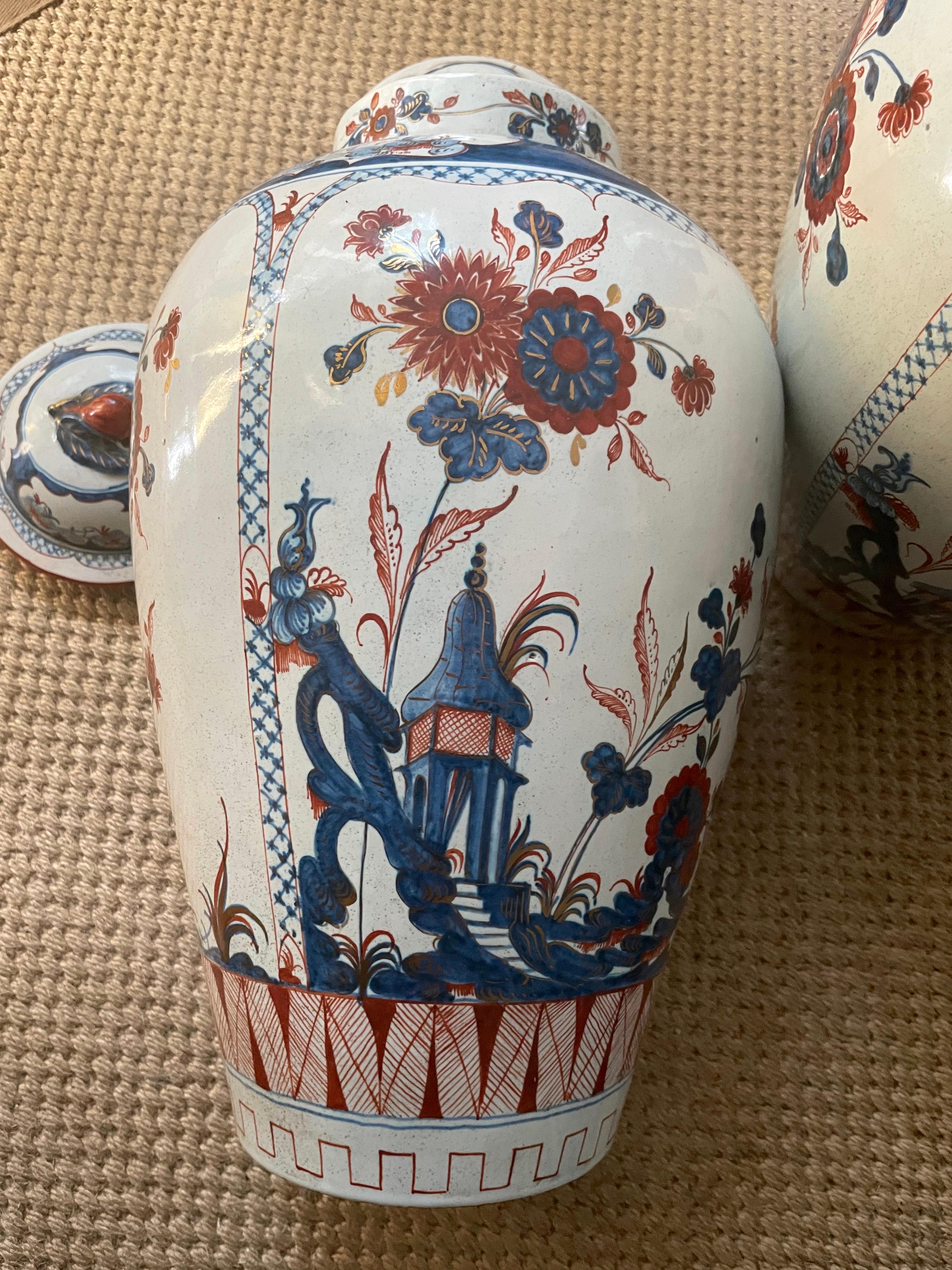 Pair blue and white chinoserie faiance vases. Pair large chinoiserie painted vases in Imari palette of blues, iron red, and gilding on cream ground with pagodas, flowers, and stylized fencing after the Doccia pattern with fruit knopfed finial lids.
