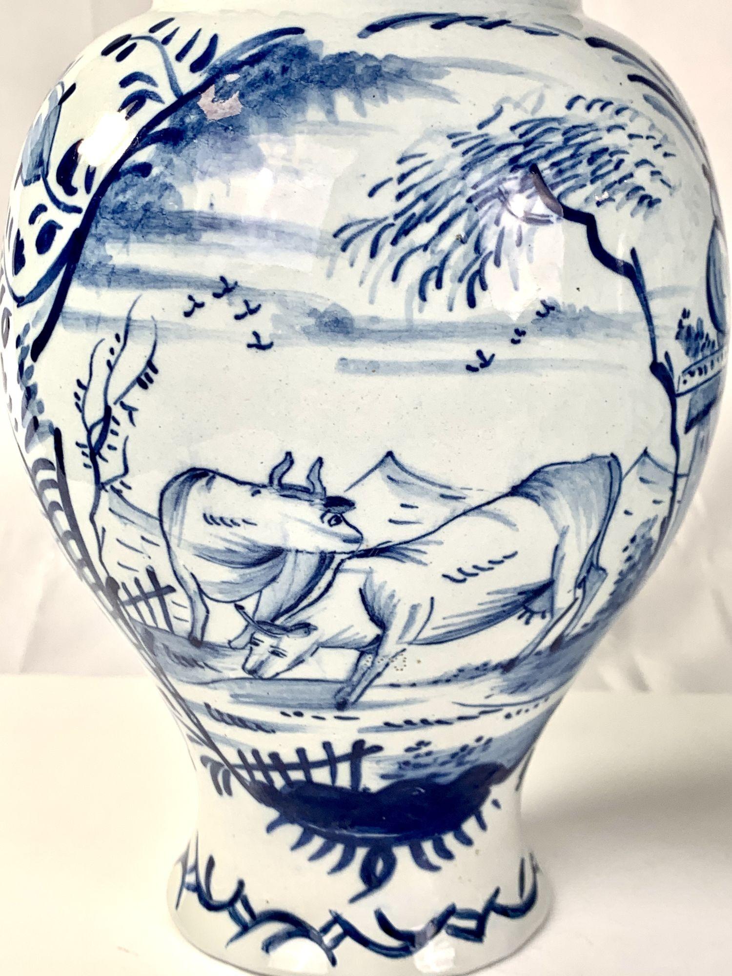 This pair of Dutch Delft mantle jars show a delightful rococo scene hand-painted on blue and white Delft.
We see a pair of cows resting in a fenced area with a flock of birds in the sky and in the background fields and mountains.
 The finial on the