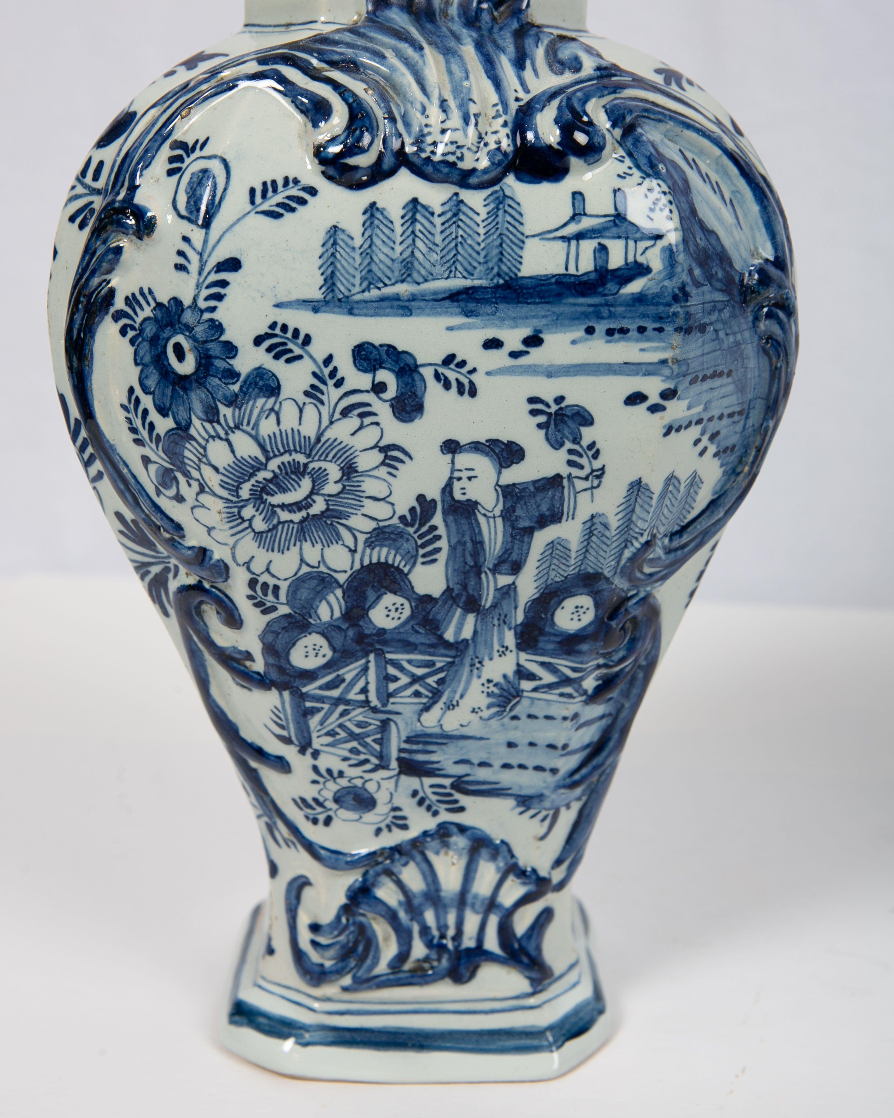 A pair of 18th-century blue and white Delft covered mantle vases hand-painted in shades of cobalt blue. The romantic chinoiserie design shows a young lady picking flowers in a garden. Around her, we see the garden fence, oversized flowers, and