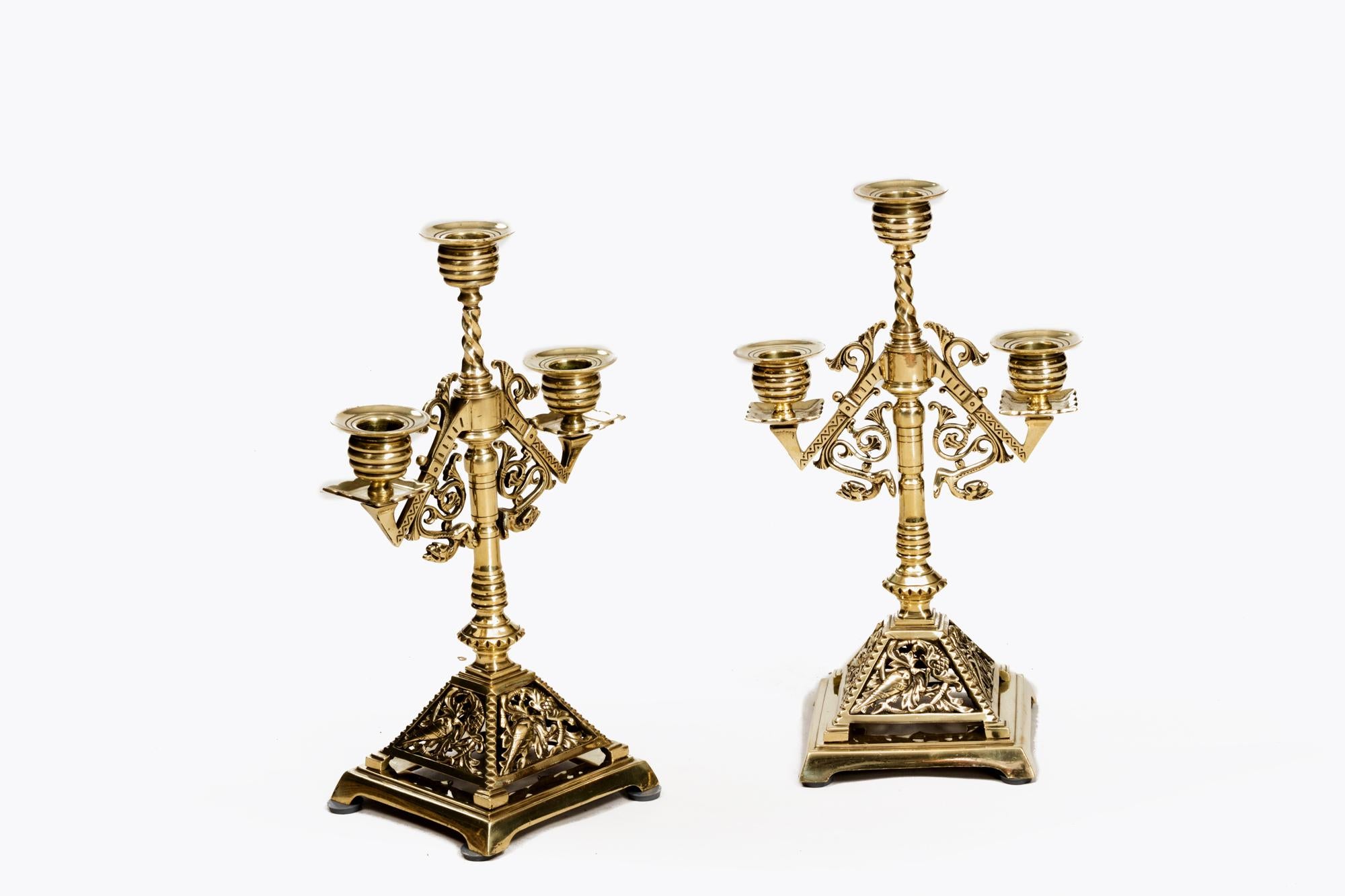 Pair brass three branch candlesticks in the Egyptianised style with acanthus leaf detailing and barley twists to the primary stems. The pair sit atop stylised pyramids with naturalistic detailing and terminate on simple square bases. 

They have