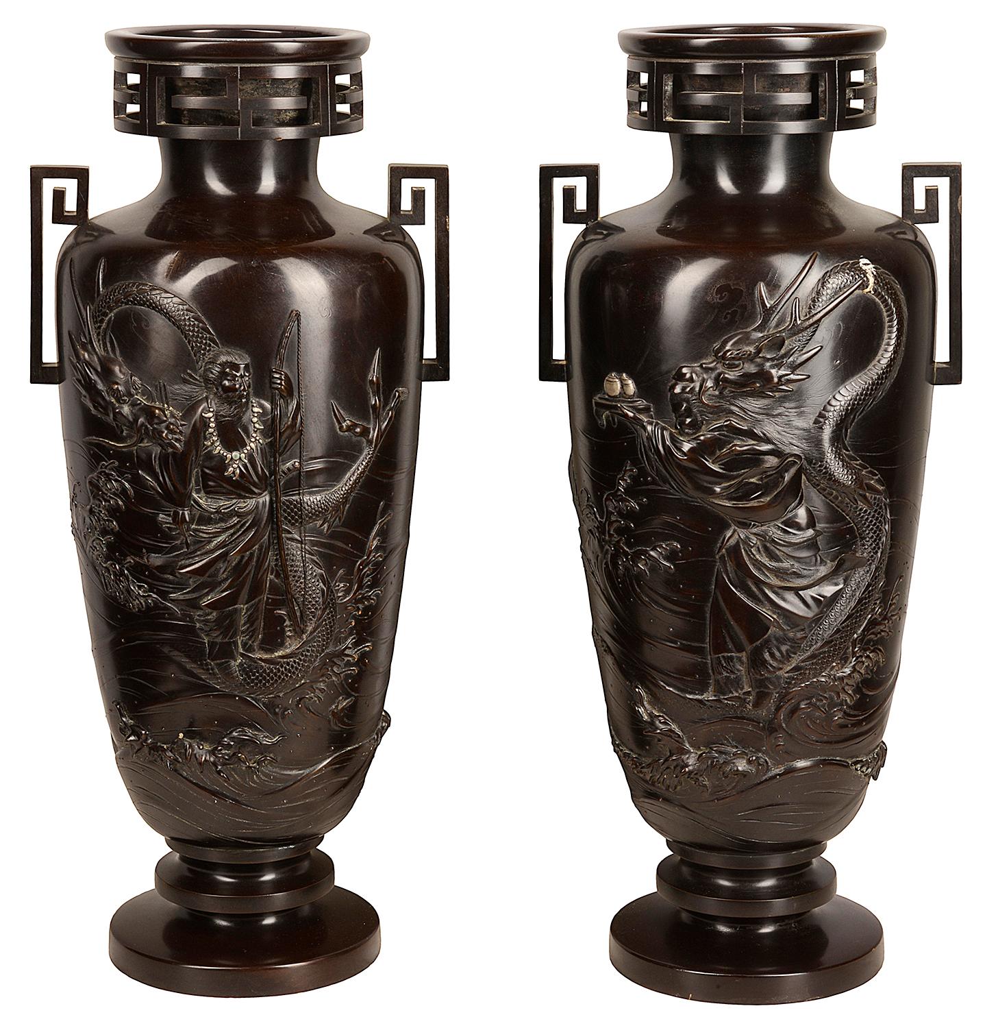 A pair of Japanese cast bronze vases, each of slender tapered form resting on a stepped base and rising to a narrow neck with everted, key-fret rim, applied to the shoulder with rinzu handles, each decorated with a mythical figure, one offering a