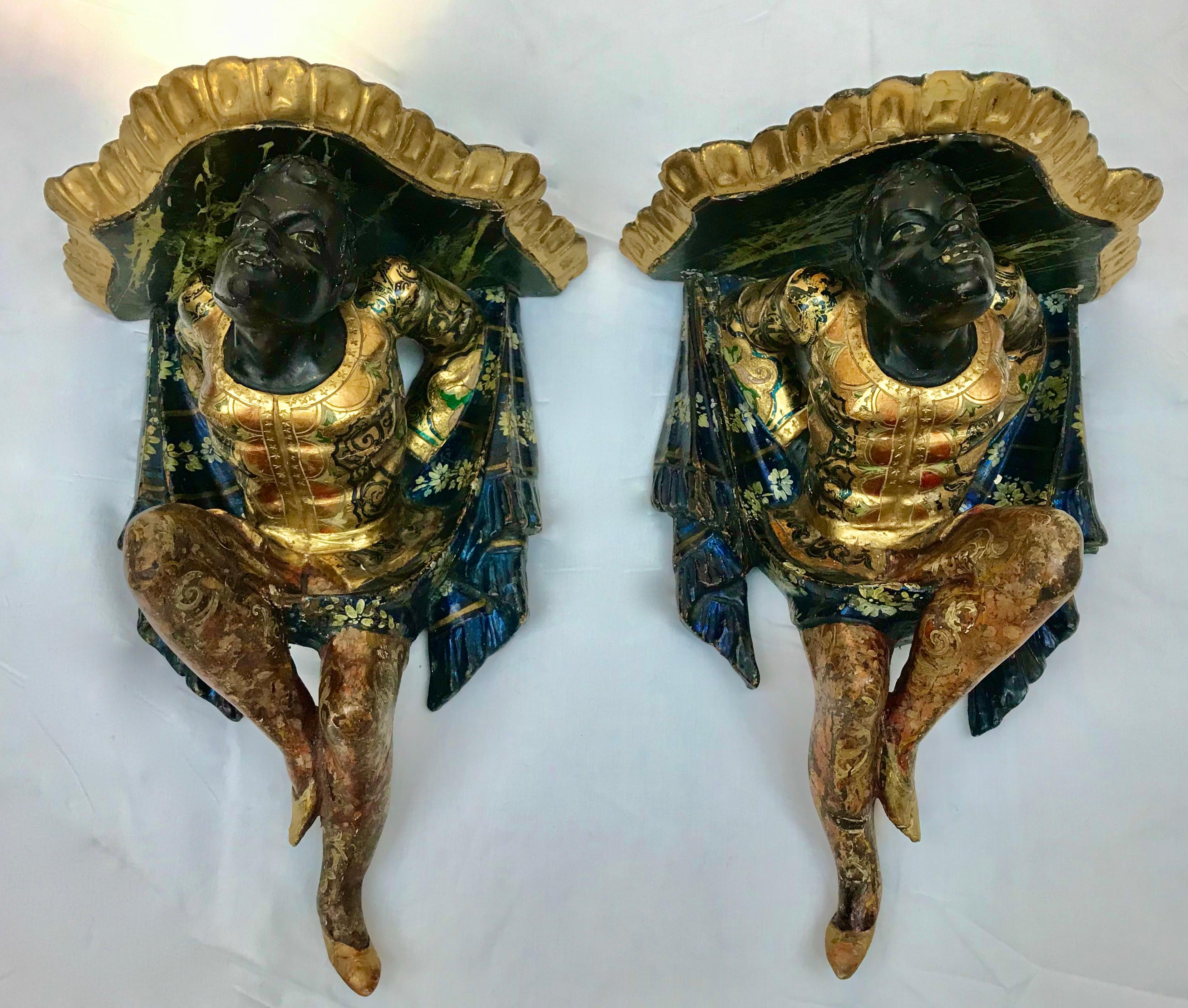 This fanciful  pair of hand carved, gilded, and polychrome figural wall brackets were made in the 19th Century in Venice, Italy.
They feature fancy dressed figures supporting faux marble shelves with carved drapery and tassels.