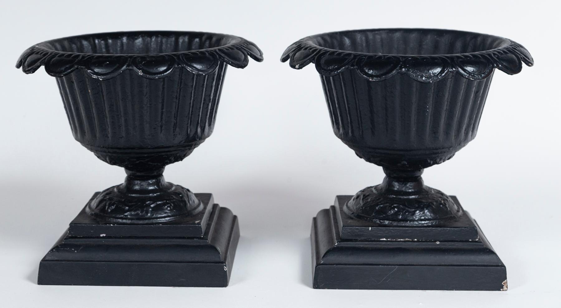 Pair of Cast Iron Urns on Bases, early 20th century. Elegant, scalloped rim with fluted body. Urns sit atop fitted bases. Weathered black painted surface.