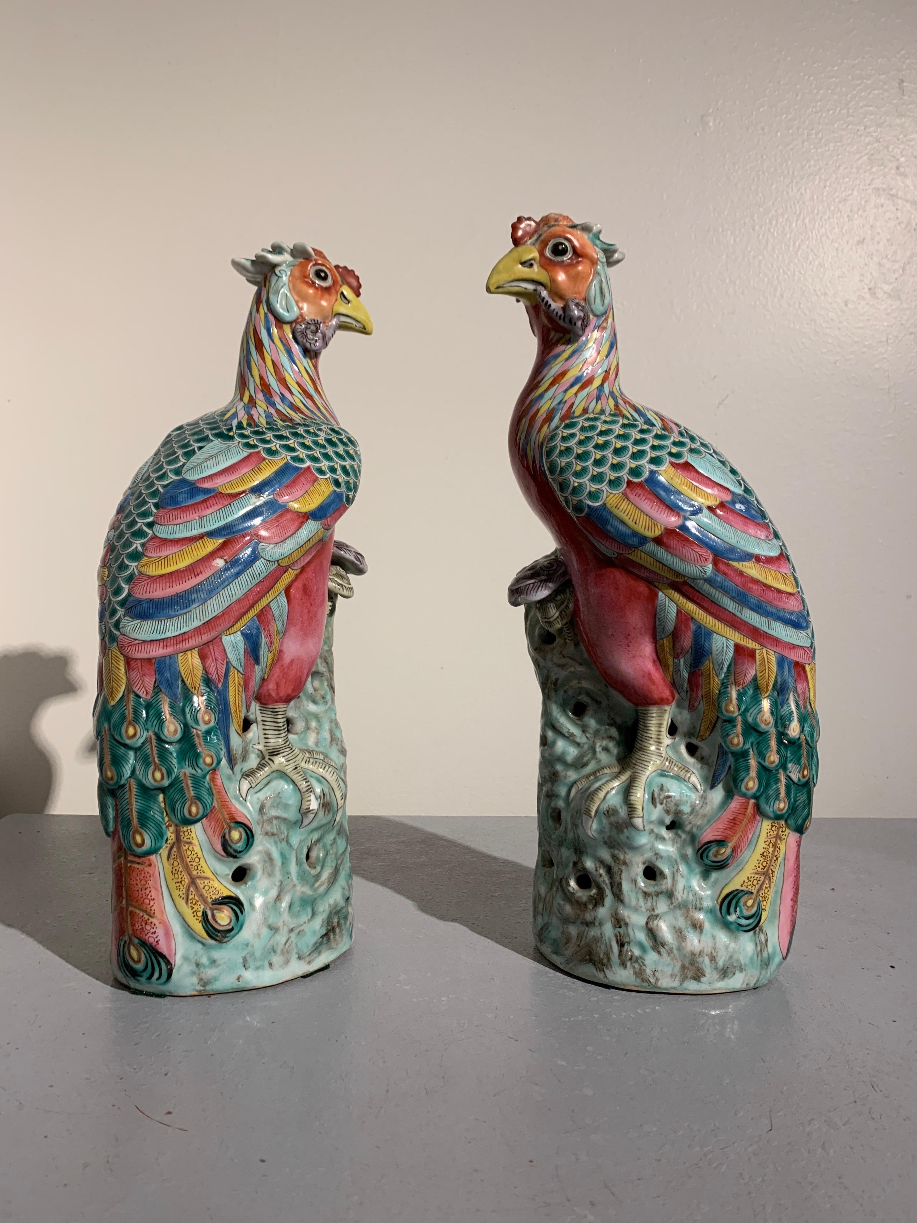 A striking pair of Chinese Republic Period famille rose enameled porcelain phoenixes, early to mid-20th century.

The majestic birds are modeled in repose, perched upon a rocky outcrop, heads turned, a spring of lingzhi (the fungus of immortality)
