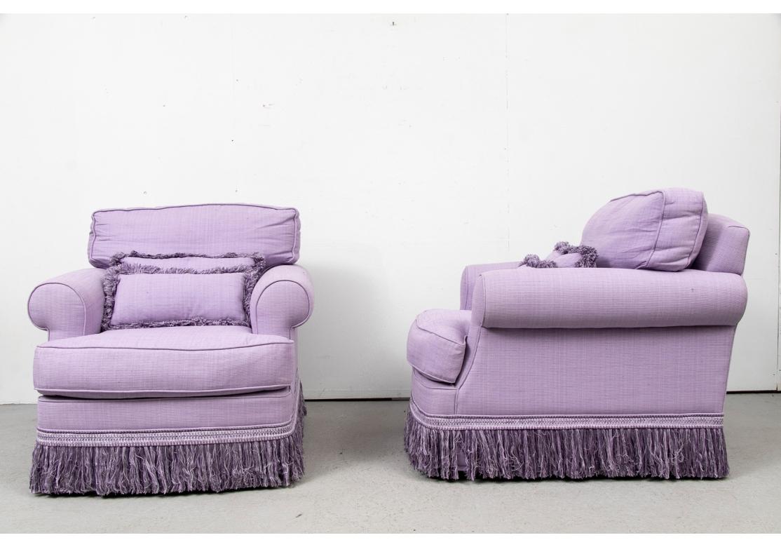 Striking pair of violet tone club chairs. Comfortable ample sized club chairs with swivel construction, rolled arms and two lumbar pillows each. Upholstered in a violet tone fabric with coordinating fringe on the skirt rails and the pillows. 
H. 32