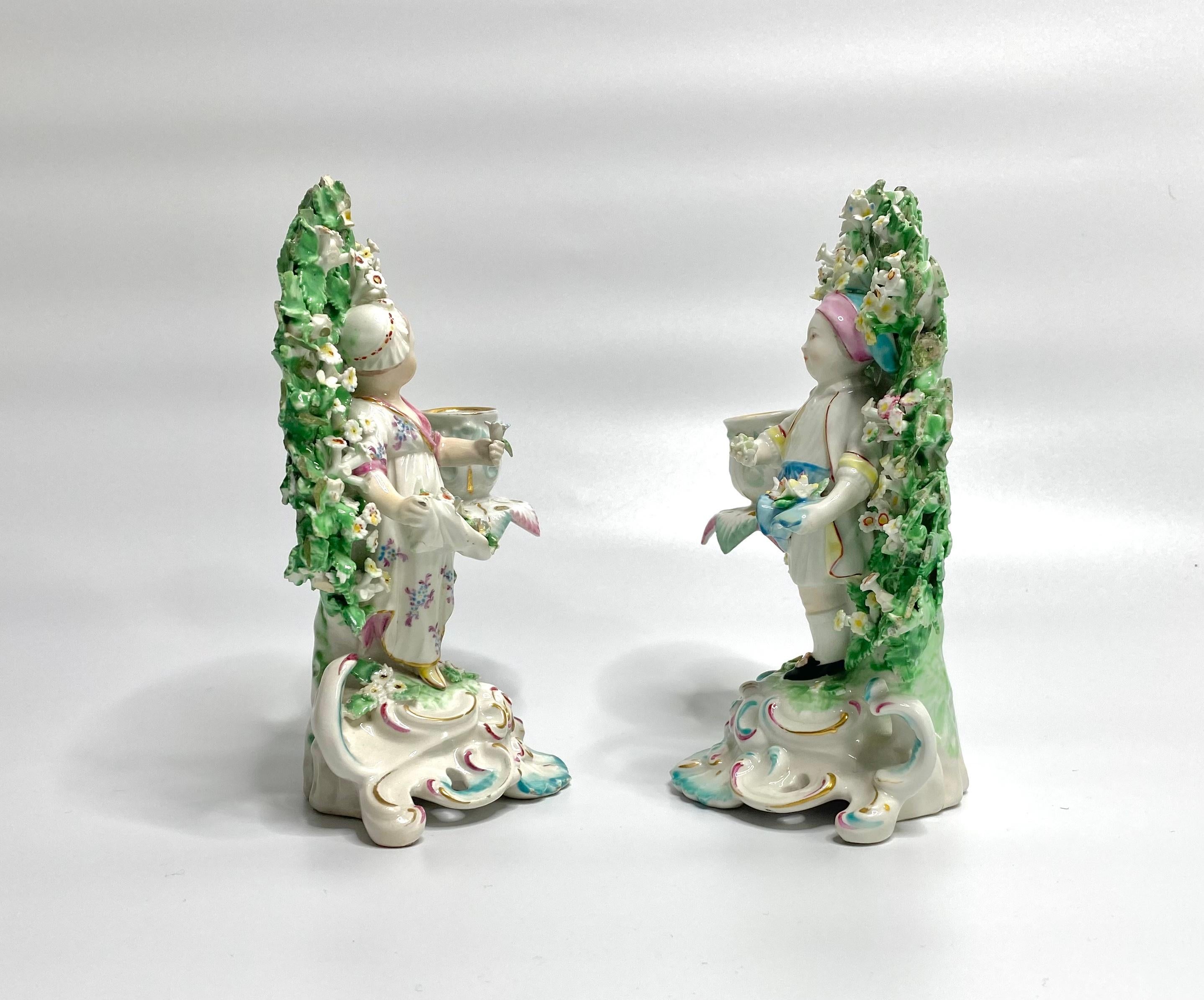 Pair of Derby porcelain bocage candlesticks, c. 1765. Modelled as a boy and girl gathering flowers in their aprons, before elaborate bocage. Each figure with a candle sconce issuing from the bocage.
Set upon elaborate rococo scroll bases, edged in