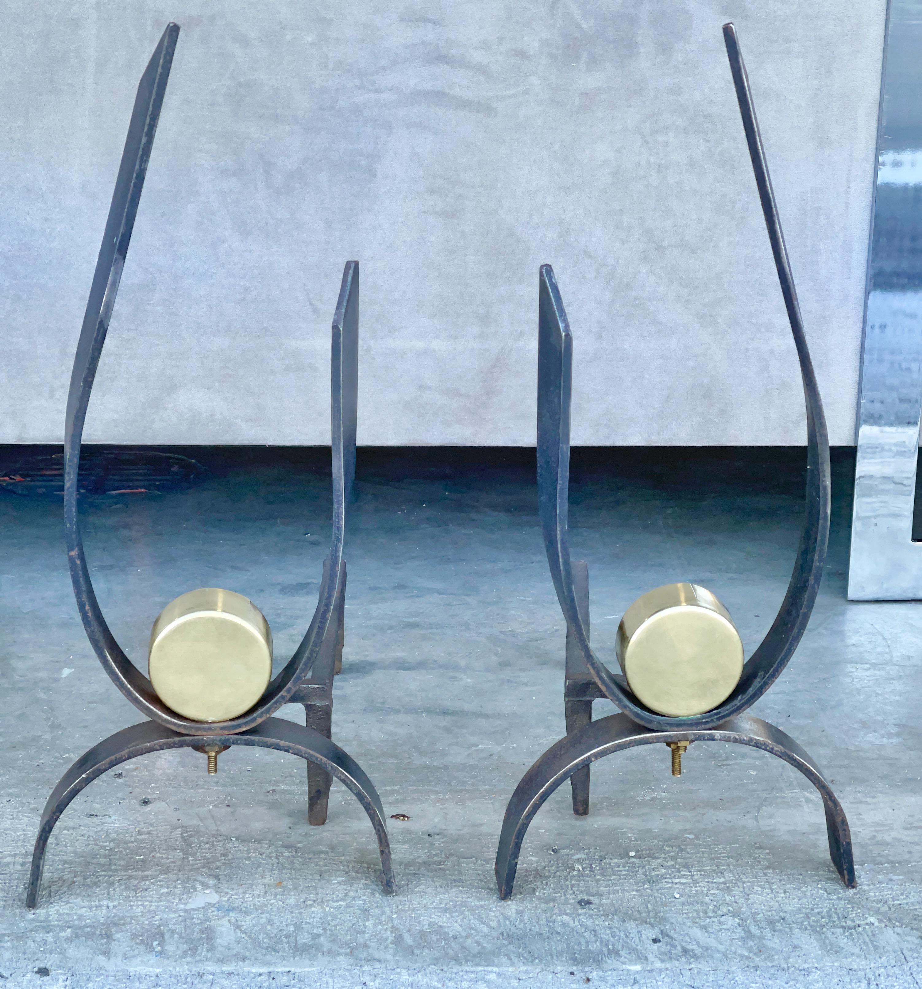 Pair of fireplace andirons designed by Donald Deskey circa 1950 for Bennett.
Sculpturally cut and formed forged blackened iron J swoops (aligned for left and right) embellished with a round brass puck sized ornaments, standing on symmetrical arched