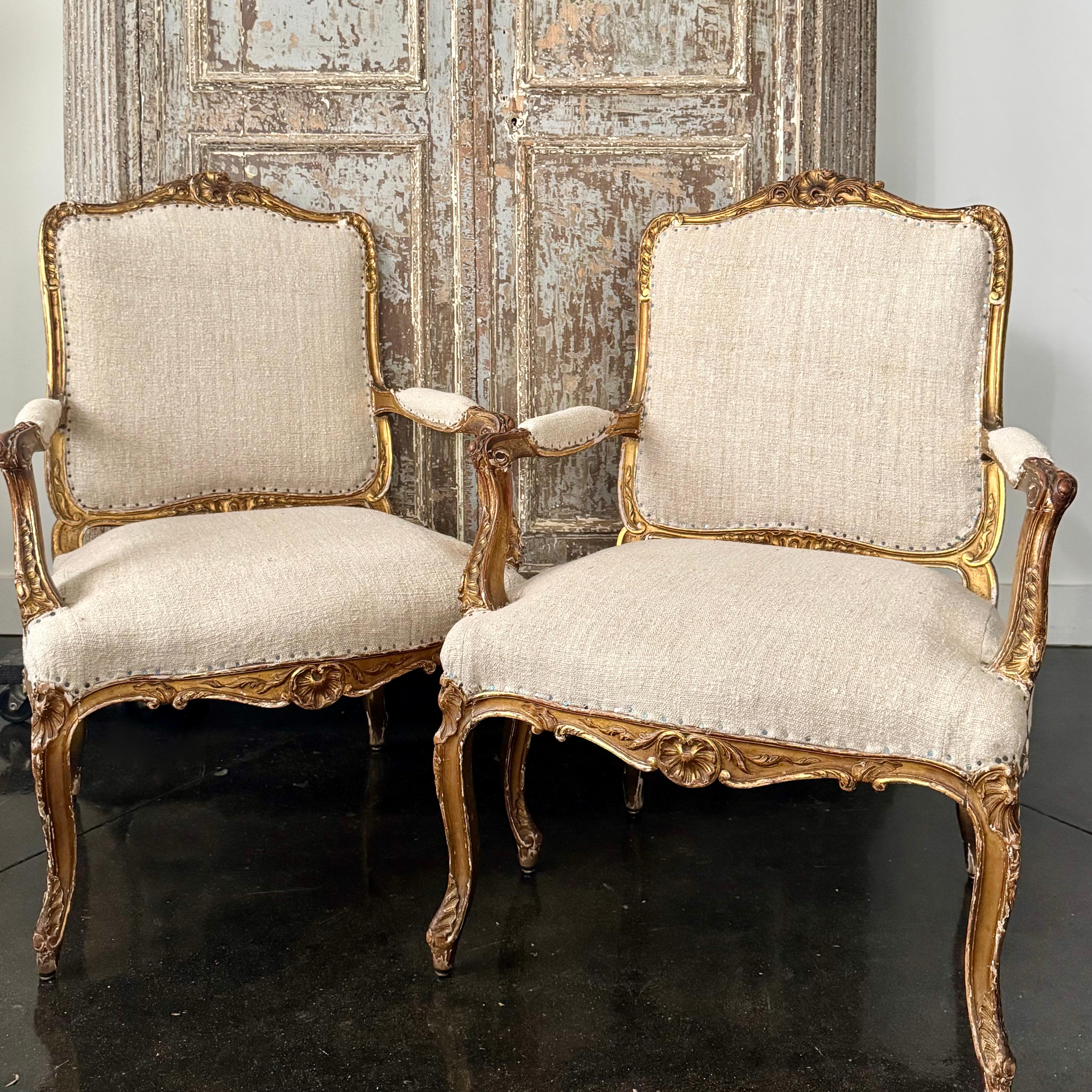 Perfectly imperfect elegant pair of 19th-century French Regency style fauteuils with  an intricately carved gilt wood frame with ornate detailing in beautiful craftsmanship with lavish floral and scroll motifs. The chairs are upholstered in natural
