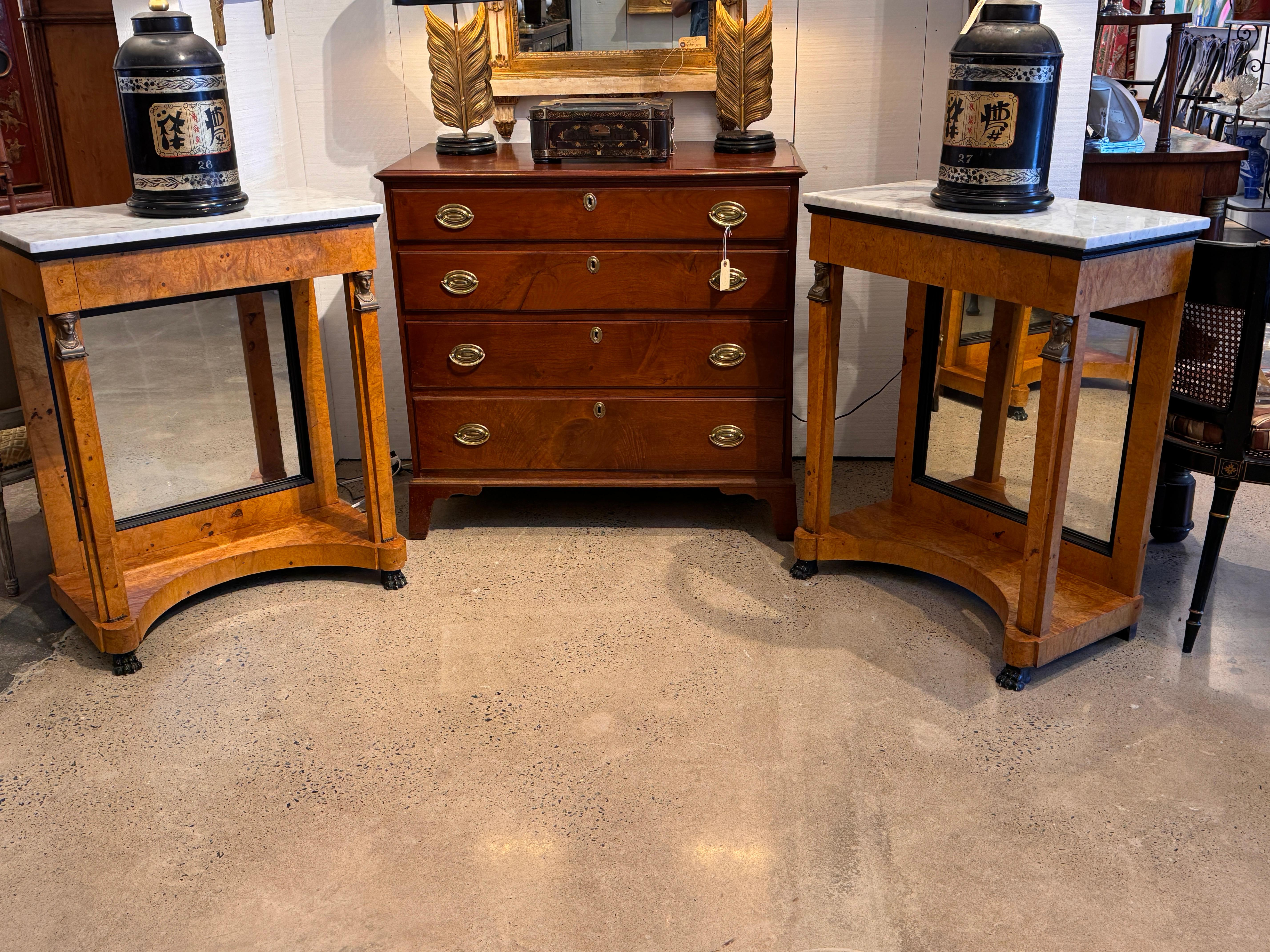 Empire-style pair of consoles with burlwood surfaces and white marble tops. Each console has a single drawer with visible dovetail joinery, mirrored backs, and fluted front supports capped with cast figural mounts. The bases are shaped with an