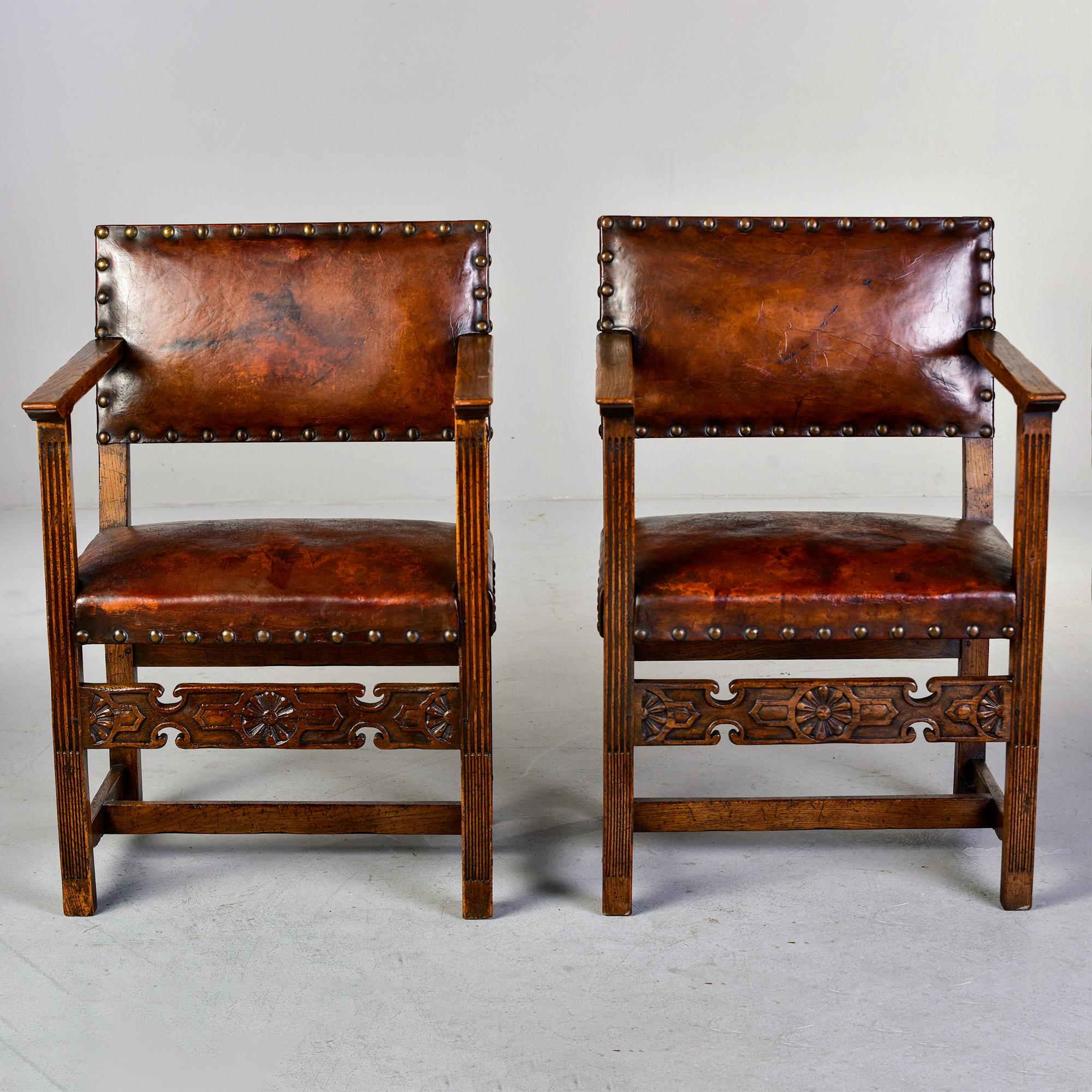 Circa 1930s pair of English oak arm chairs in Arts and Crafts style. Chairs feature carved details on stretchers, original leather affixed with large brass head tacks.

Very good antique condition with original leather and wood showing honest