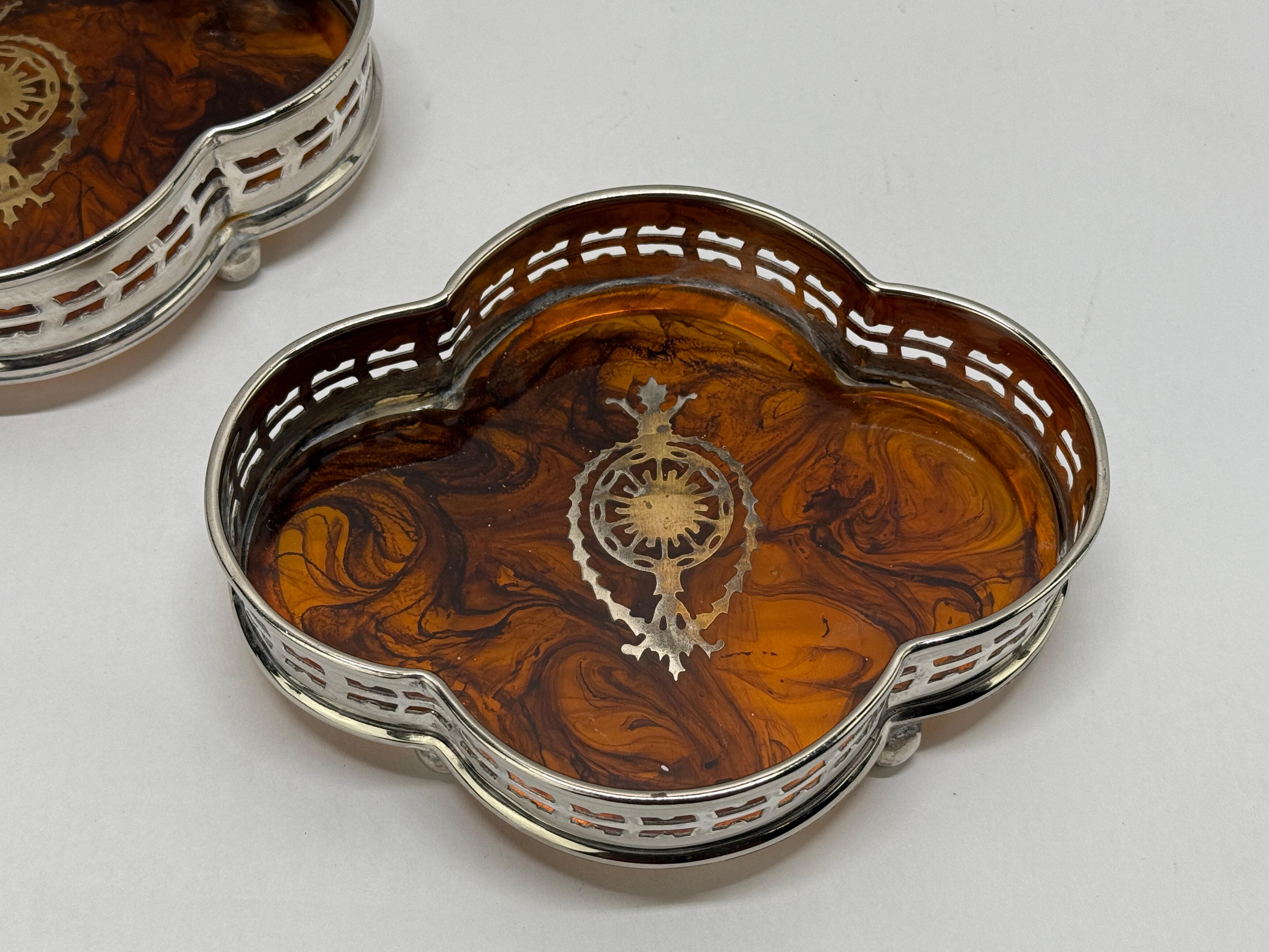 Cette élégante paire de sous-verres à vin vintage illustre le savoir-faire raffiné et le design sophistiqué typiques de la vaisselle décorative du début et du milieu du XXe siècle. Fabriqué en métal argenté brillant, chaque sous-verre présente un