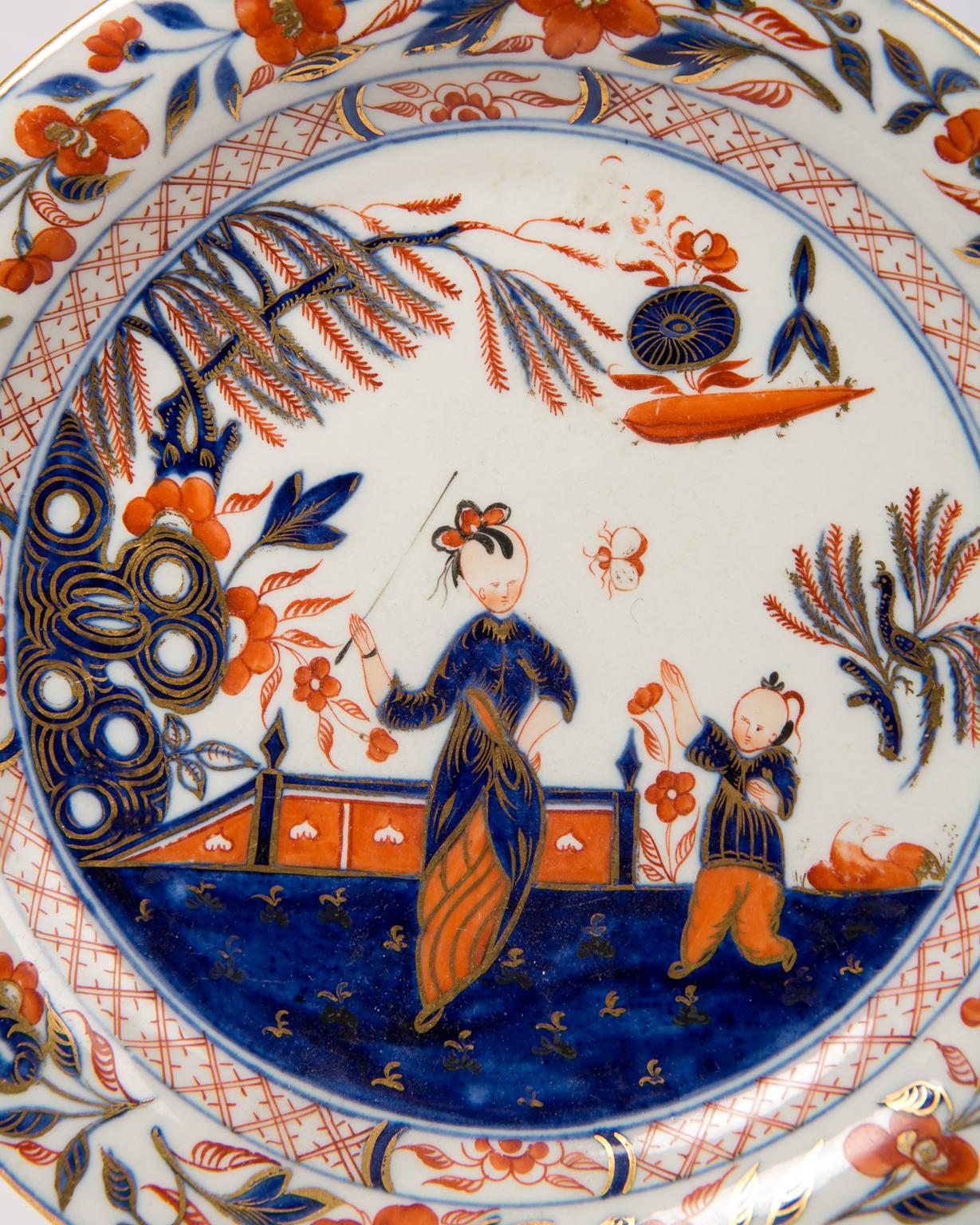 WHY WE LOVE IT: The colors! The scene!
We are pleased to offer this pair of English dishes inspired by Imari decoration showing a scene painted in cobalt blue, red and gold. We see a lady walking in a garden with her young son. The branches of a