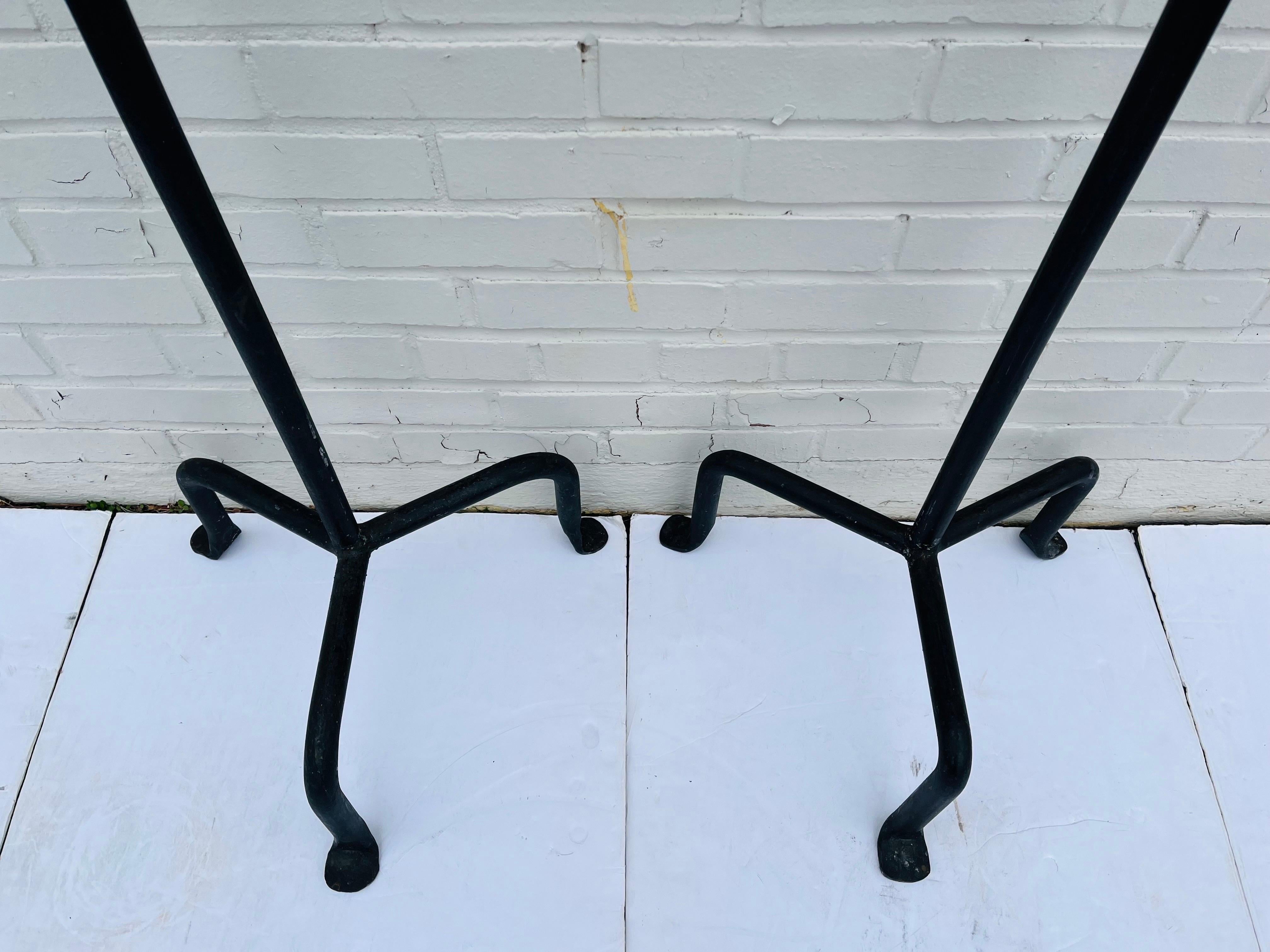A vintage pair of wrought iron candle stick torchieres in the Post Modern style. Each floor candle stick features a tripod base with splayed, wrought feet hammered to a rounded fan shape. The shaft of the candle stick has a looping bend somewhat in