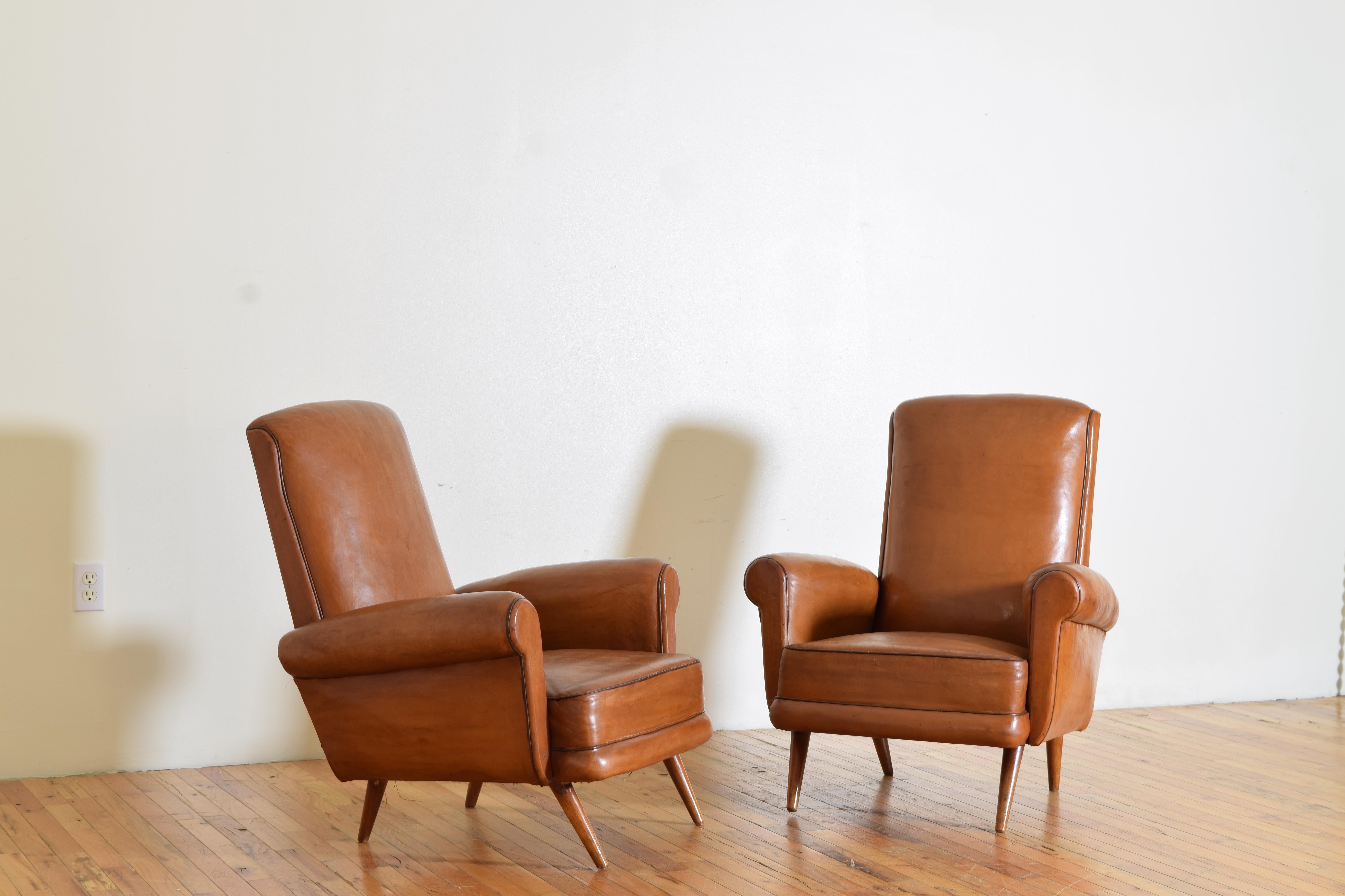 Covered in original unblemished leather with slightly darker leather piping, having rounded backs and closed rounded arms with tight seats, raised on circular tapering splayed walnut legs