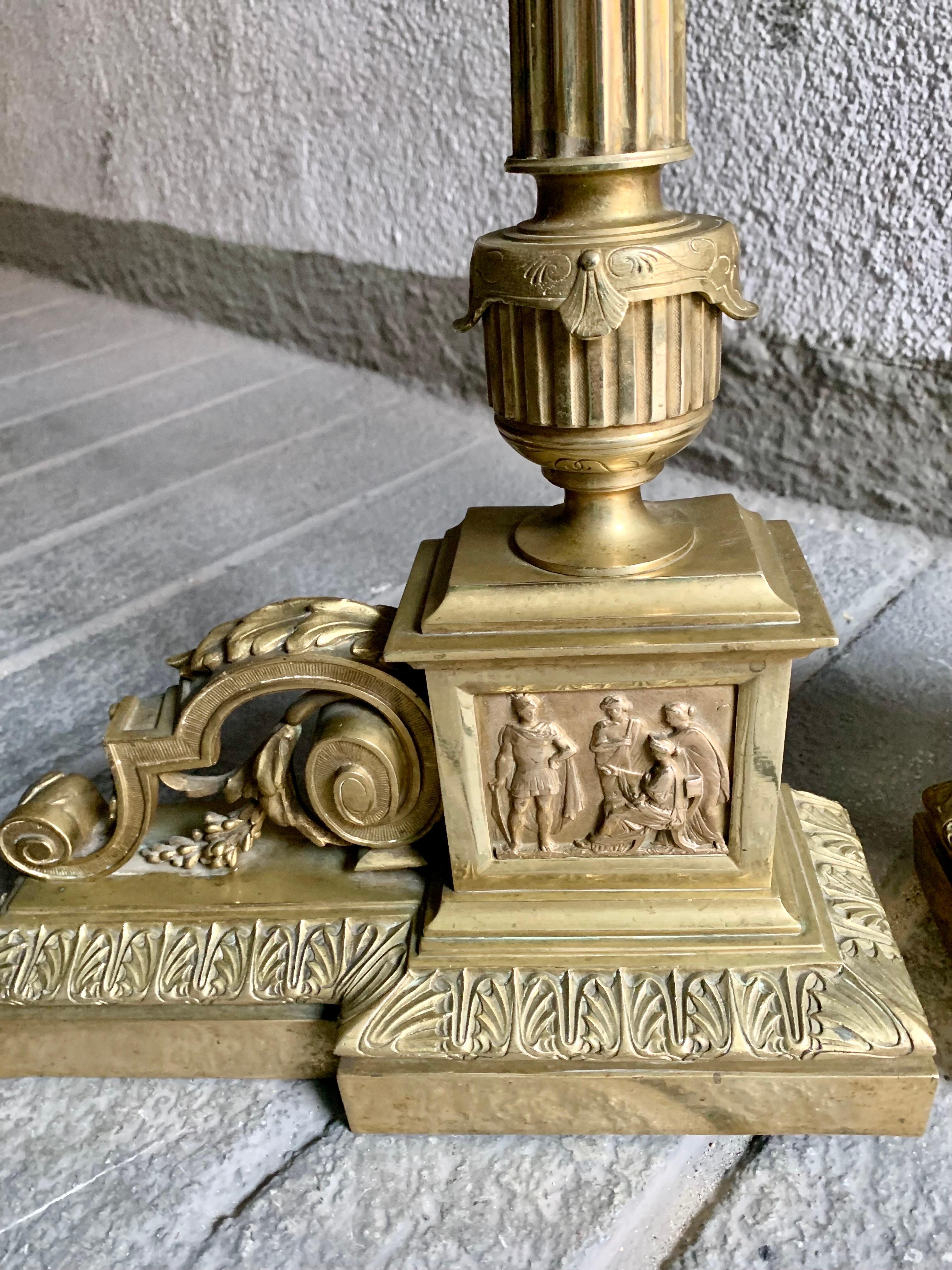 A pair of large empire-style fireplace andirons, in gilded brass, they are formed by long columns that rest on a base with a mythological scene, the central column ends in an imitation of a roman-style oil lamp, it has lateral decoration of scrolls