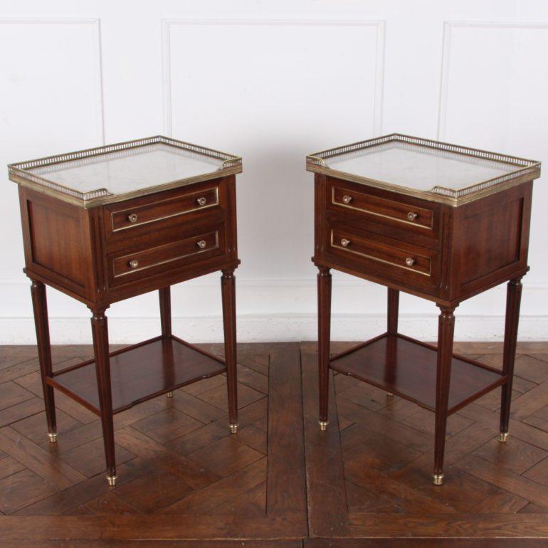 A pair of French Directoire-style mahogany bedside, or side, tables, each with a Carrara marble top and pierced brass gallery, and two drawers accented with brass trim. They are supported on turned tapering fluted legs and feature a lower shelf for