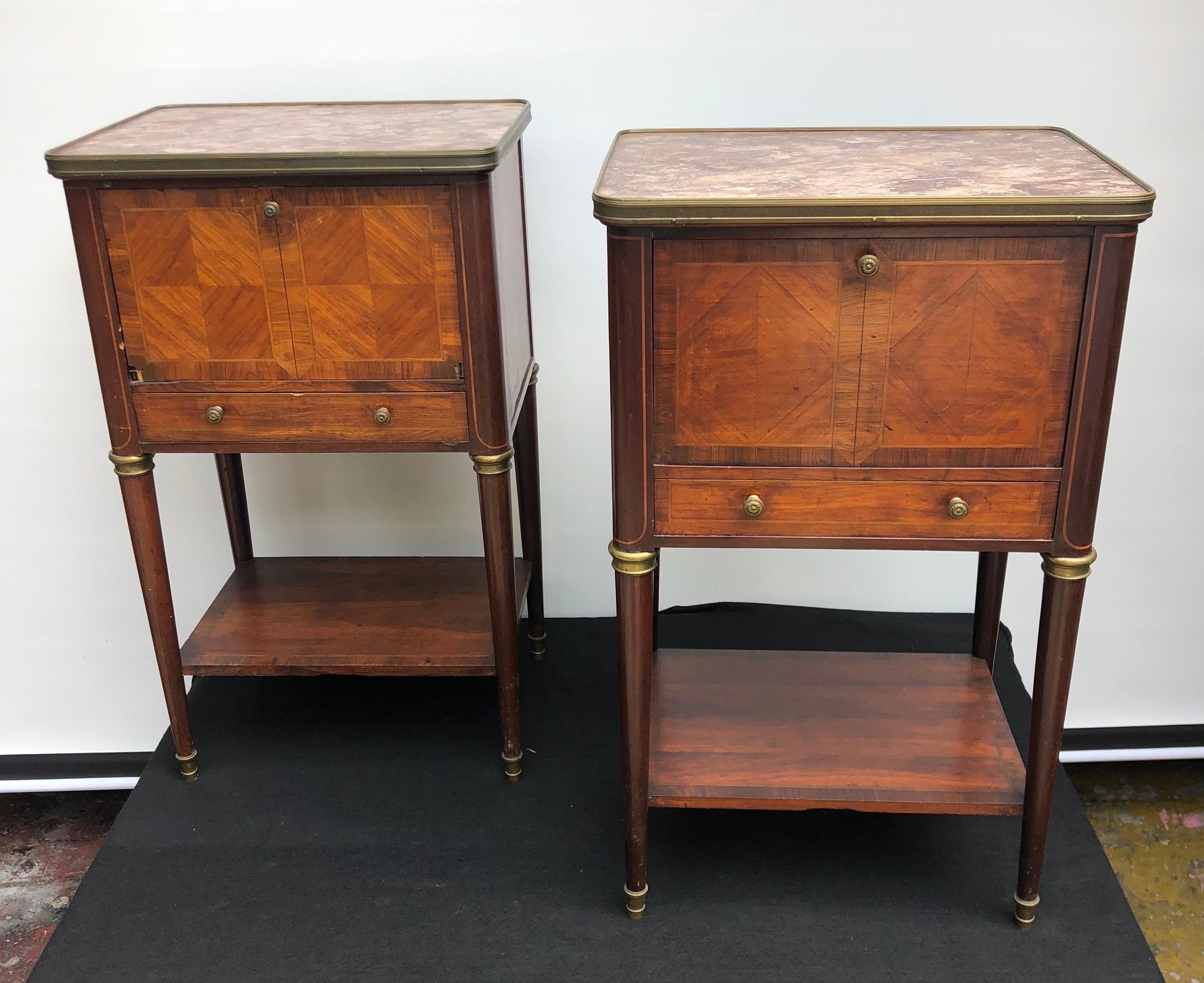 A stately pair of French Directoire style marble-top end tables / nightstands made with Mahogany, Kingwood Crossband, Satinwood inlay mounted with Bronze having a drawer with a cupboard door above covering a porcelain lined interior.