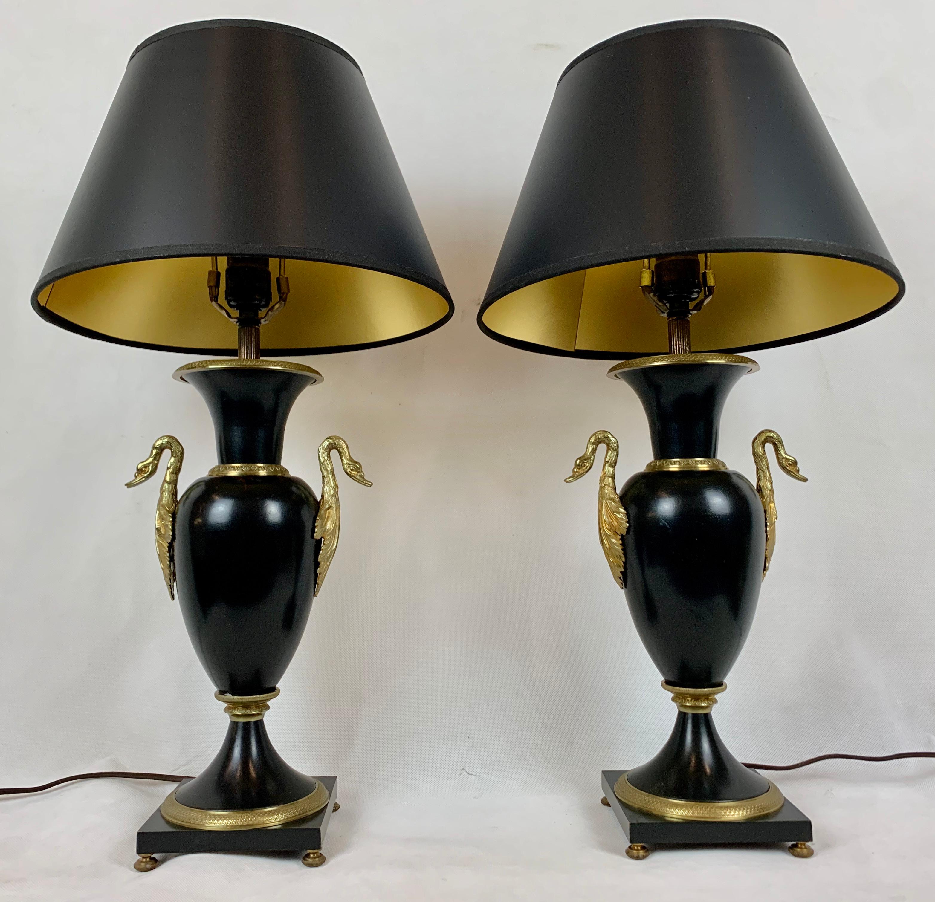 Pair of French Empire black enameled lamps with gilt bronze trim. The vase shaped lamps have swan handles and they sit on square bases with small squashed bun feet. 
The gilt bronze trim is finely chased (see photos). The black hard back paper