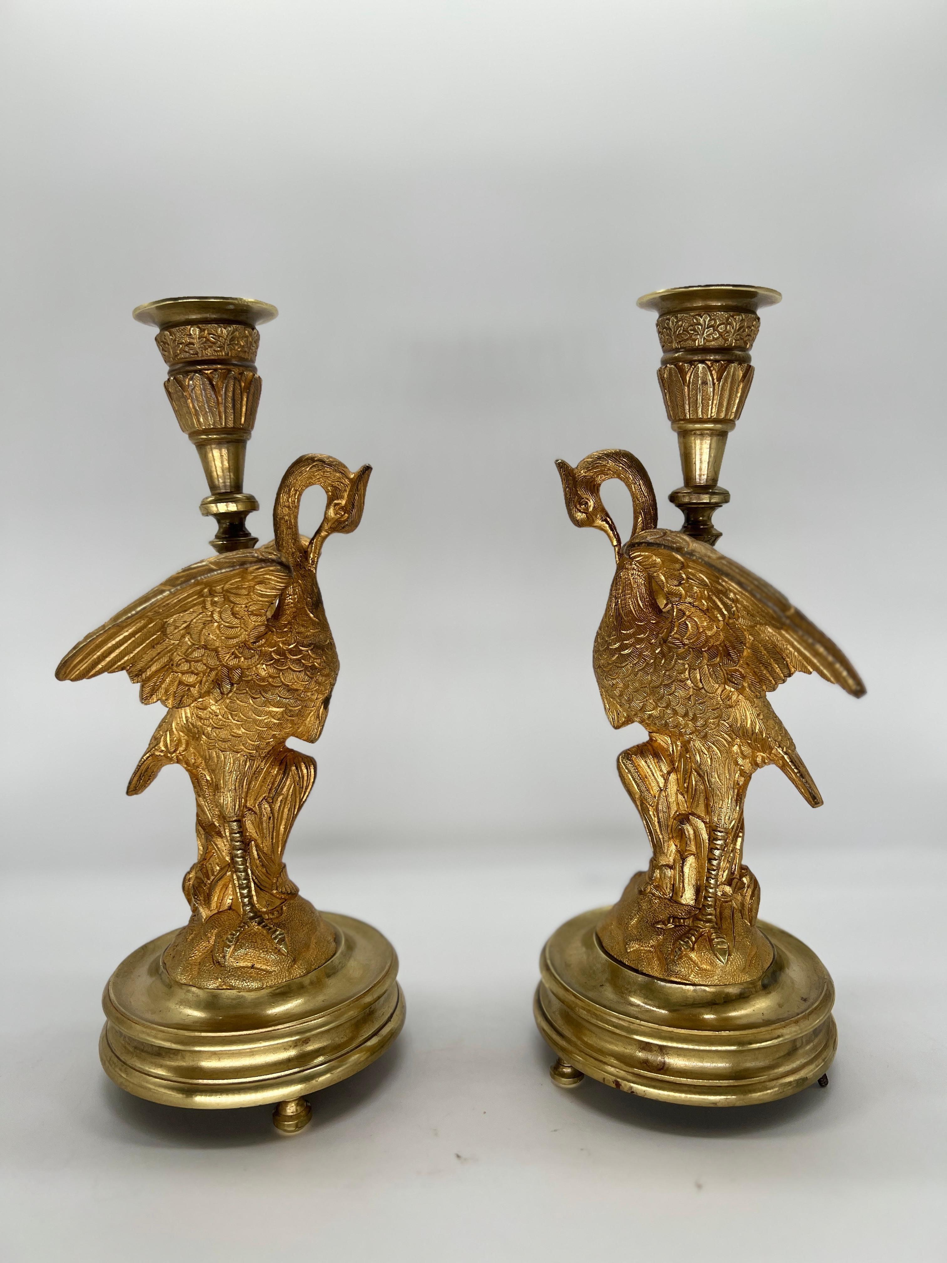 French, circa 1815. 

This exquisite pair of French Empire gilt bronze candlesticks, crafted circa 1815, embodies the opulence and grandeur of the Empire style. Each candlestick features a graceful crane motif, symbolizing longevity and prosperity,