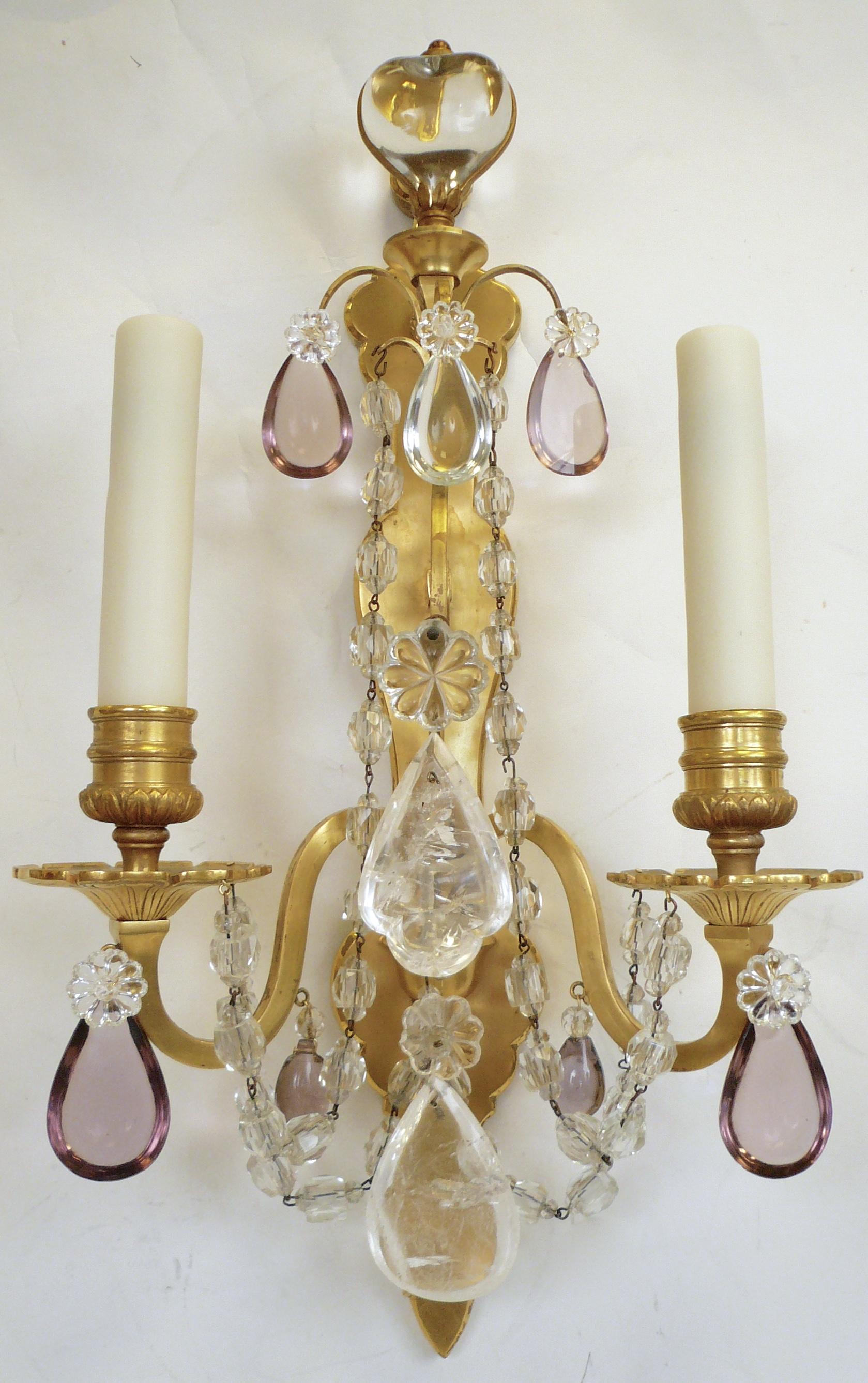 This beautiful pair of Louis XVI style twin arm sconces feature neoclassical motifs including acanthus leaves. They are trimmed in clear and amethyst cut crystal prisms, and rock crystal pendants.