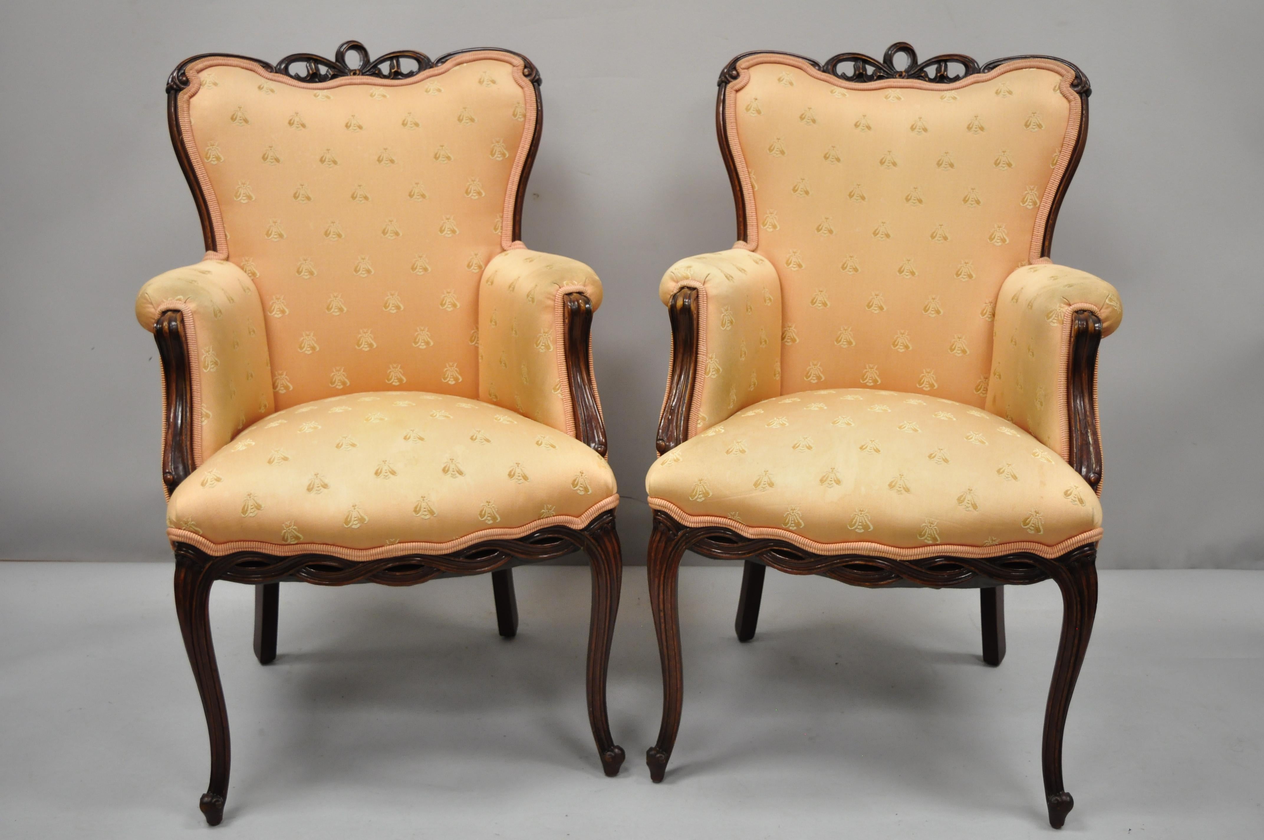 Pair of vintage French Hollywood Regency Victorian style upholstered fireside lounge armchairs. Items feature ribbon / bow carved upper rail, pretzel carved lower rail, solid wood frame, upholstered armrests, cabriole legs, mid-20th century.