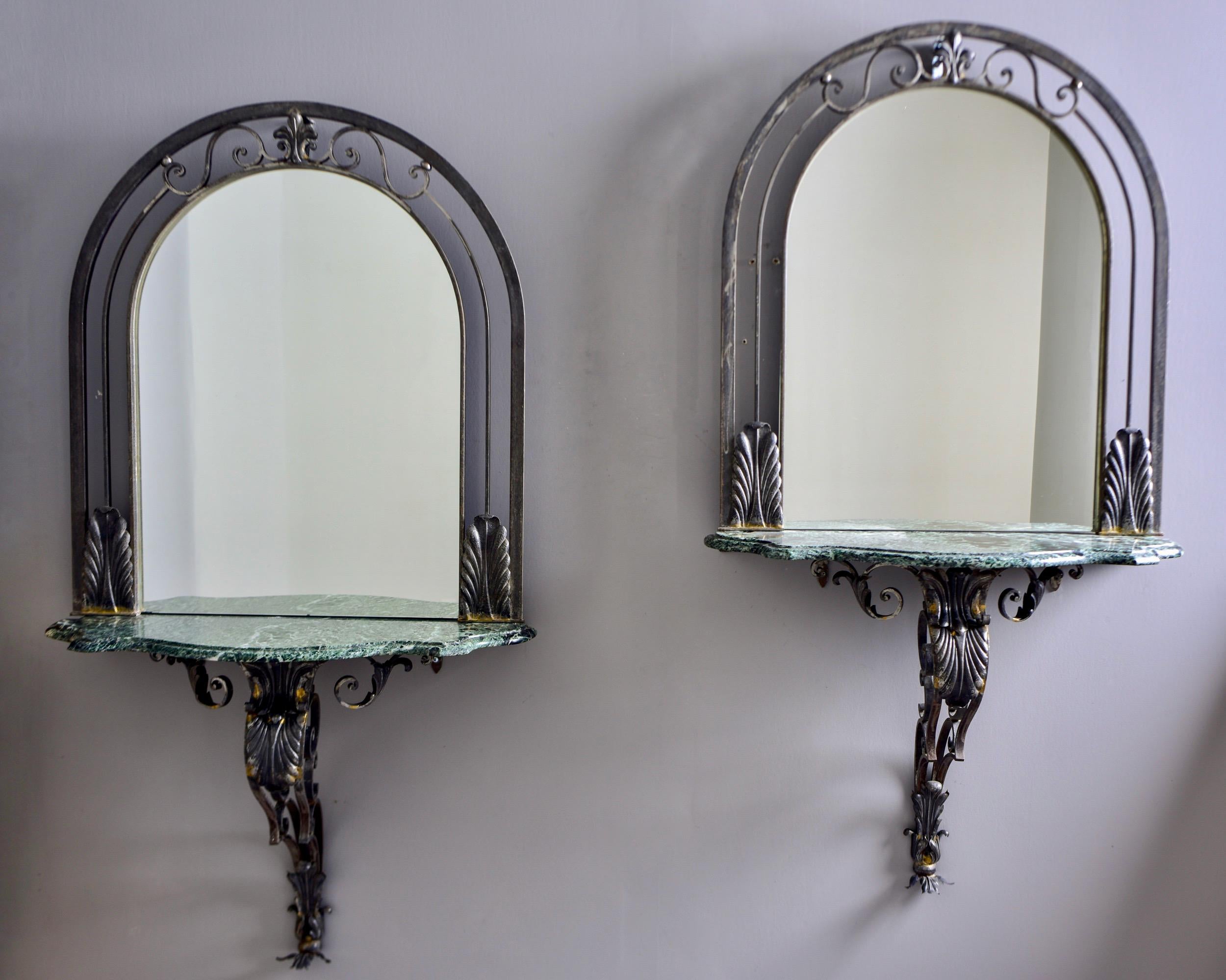 Circa 1930s pair of French wall consoles with iron-framed mirrors. Wall mounted arched iron frames feature open work and metal tole details, green marble table tops and decorative iron supports. Sold and priced as a pair. Unknown maker.