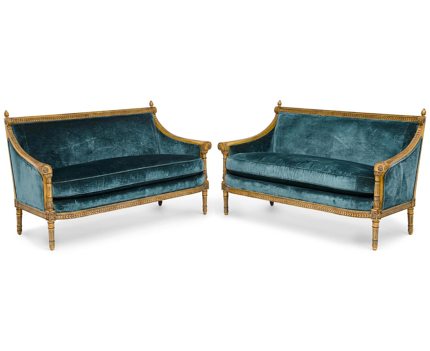 A pair of Louis XVI style carved and Laqué gilded bergère settees. The elegant pair of carved loveseats, upholstered in a blue velvet, with padded backs and sides and a cushion seat, the swagged armrests decorated with floral rosettes and topped