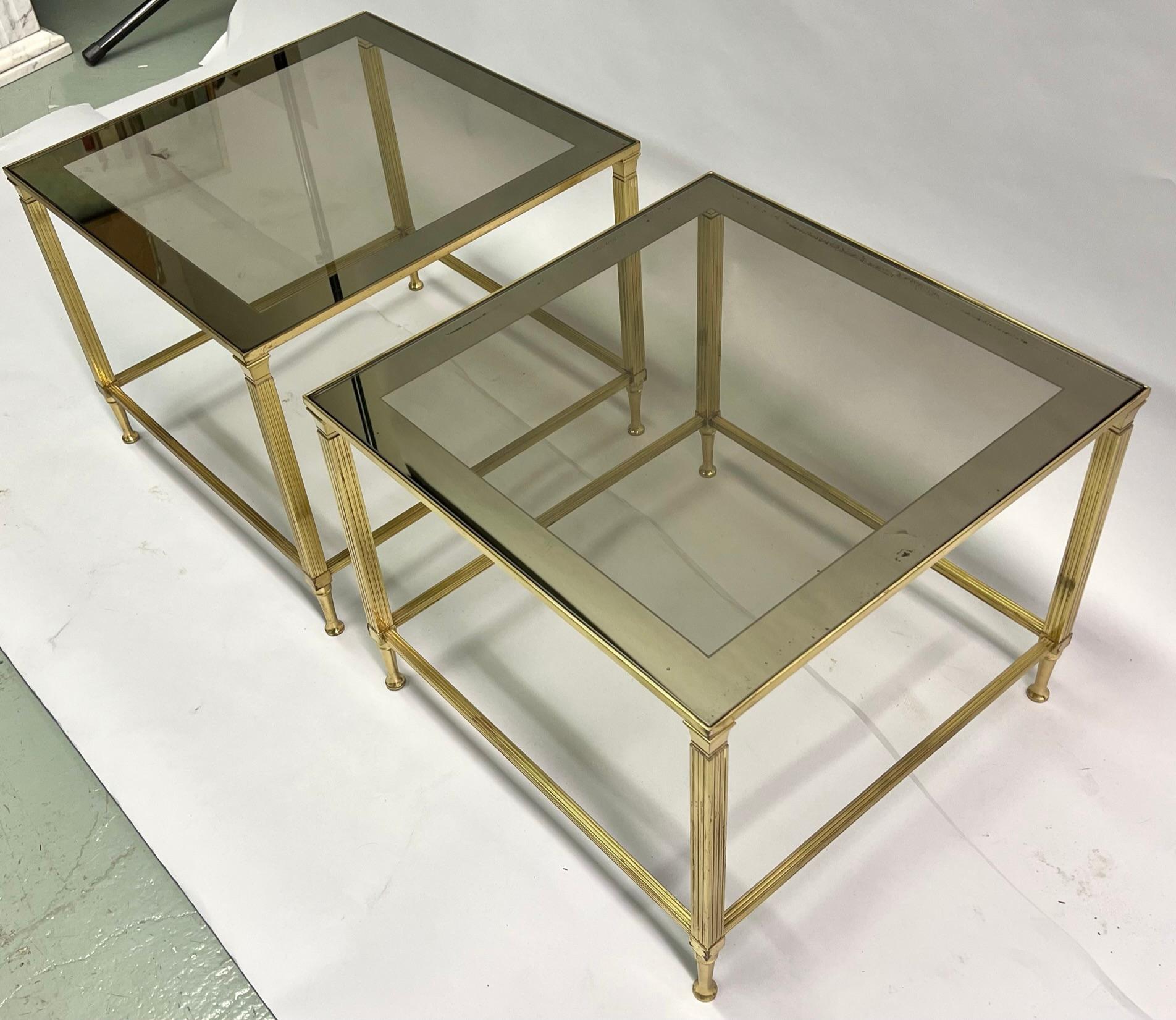 Pair of French Mid-Century brass end or side tables in the modern neoclassical style attributed to Maison Jansen. The pieces are constructed in brass and have delicate fluted pilaster legs and tapered sabots. The tops are composed of smoked glass