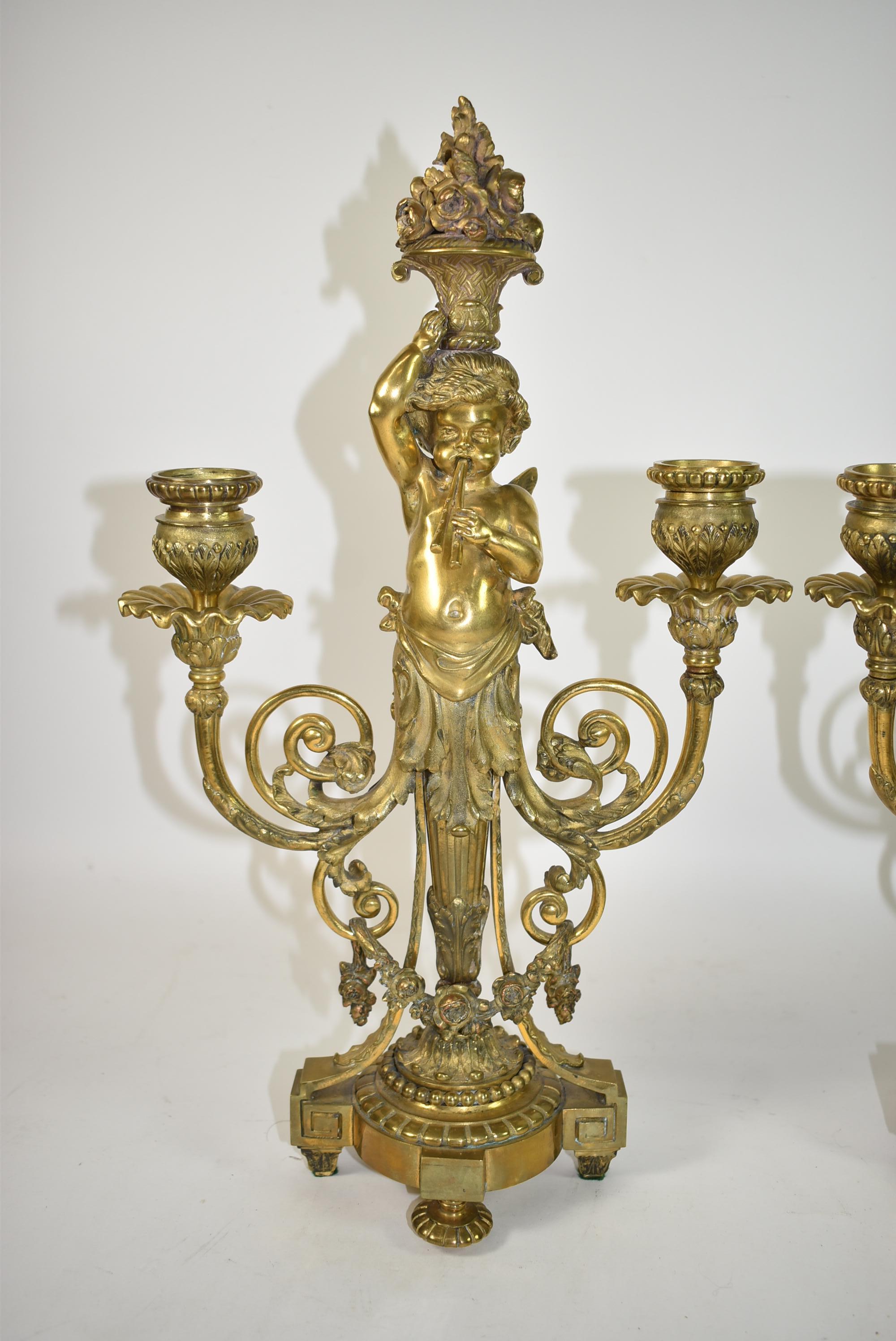 Pair of French neoclassic gilt bronze Putti cherub candelabras circa 1910. Acanthus leaf details with rose swags, scrolls and ruffled candleholders. Cherub Putti central figures hold baskets of flowers on the their heads playing double flute