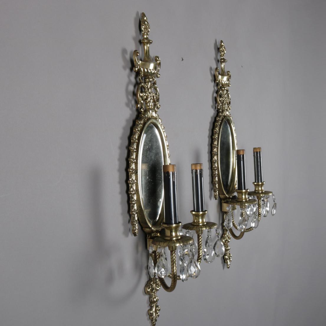 A pair of French neoclassical wall sconces feature cast brass frames having urn form crests surmounting bevelled mirrors flanked by rope and tassel form frame and over double electric candle lights with hanging cut rock crystals, 20th