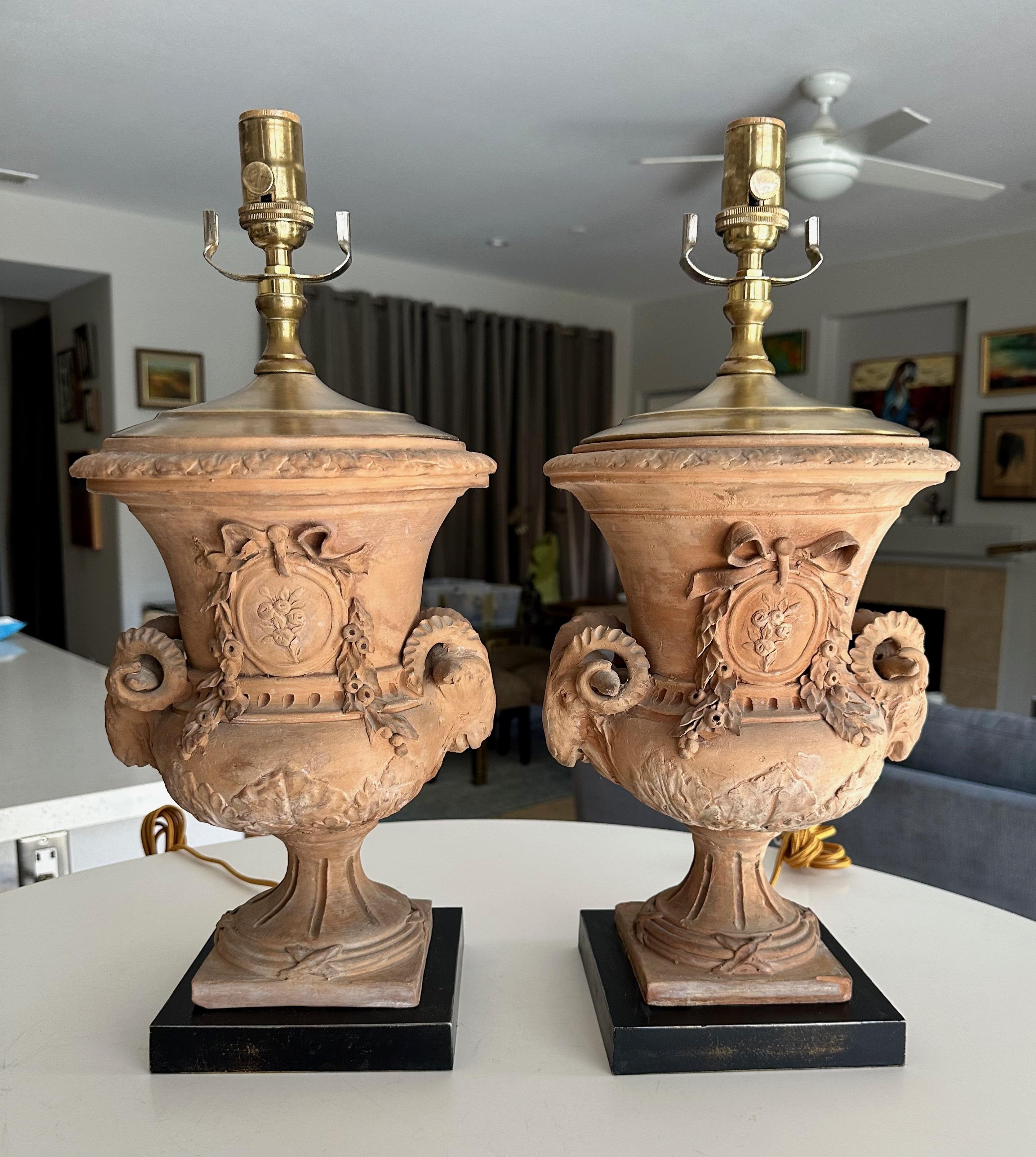 Coppia di urne in terracotta francese in stile neoclassico trasformate in lampade da tavolo. Le urne sono decorate con teste di montone a 2 manici e piaghe floreali con nastri e festoni. Le urne poggiano su basi in metallo laccato nero invecchiato,