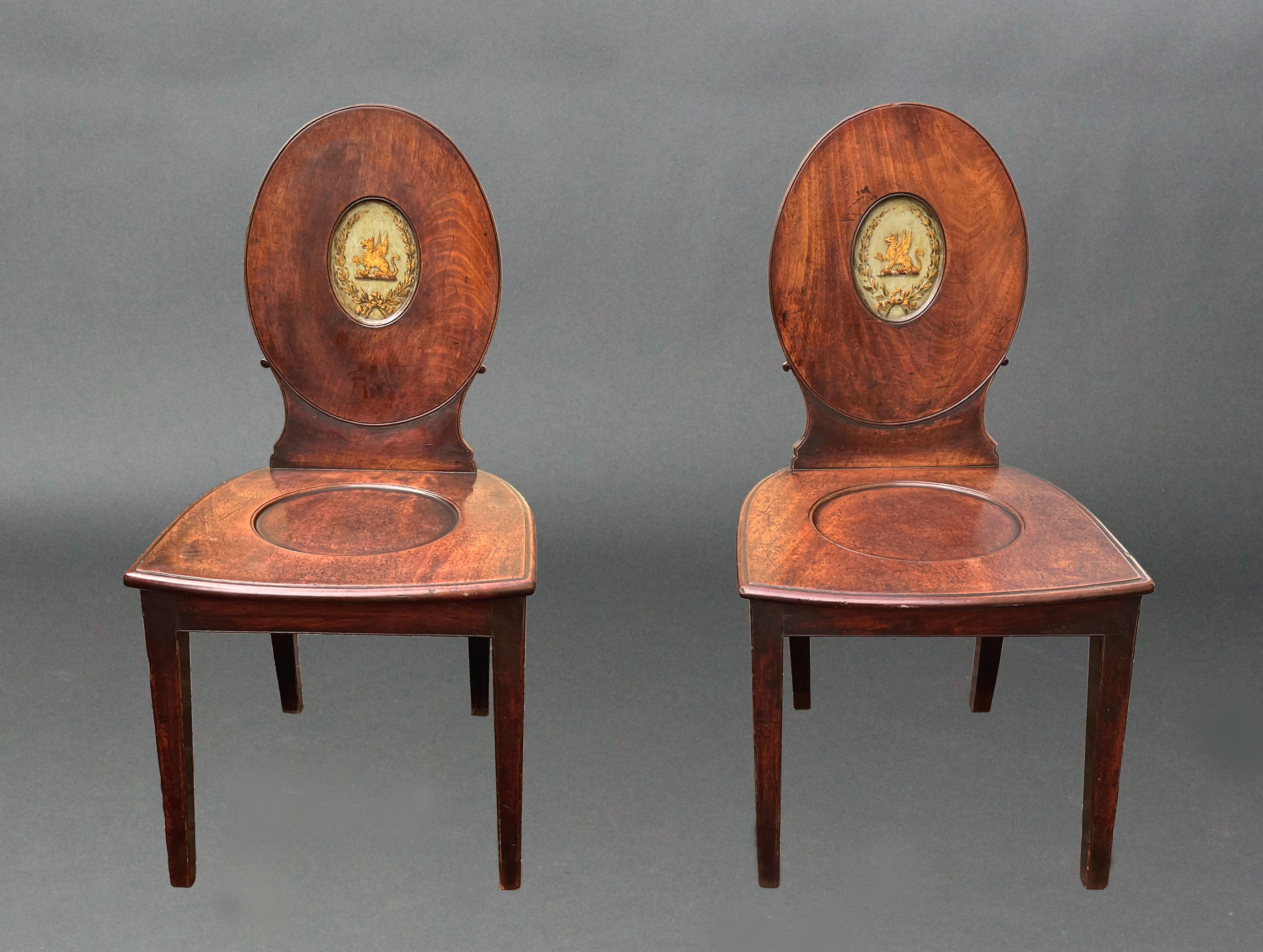 A pair of George III Hepplewhite period hall chairs of a good original colour and patina; the oval backs with centres finely decorated with gilded griffins and laurel wreaths on a pale green ground. The wide bow fronted dished seats raised on square