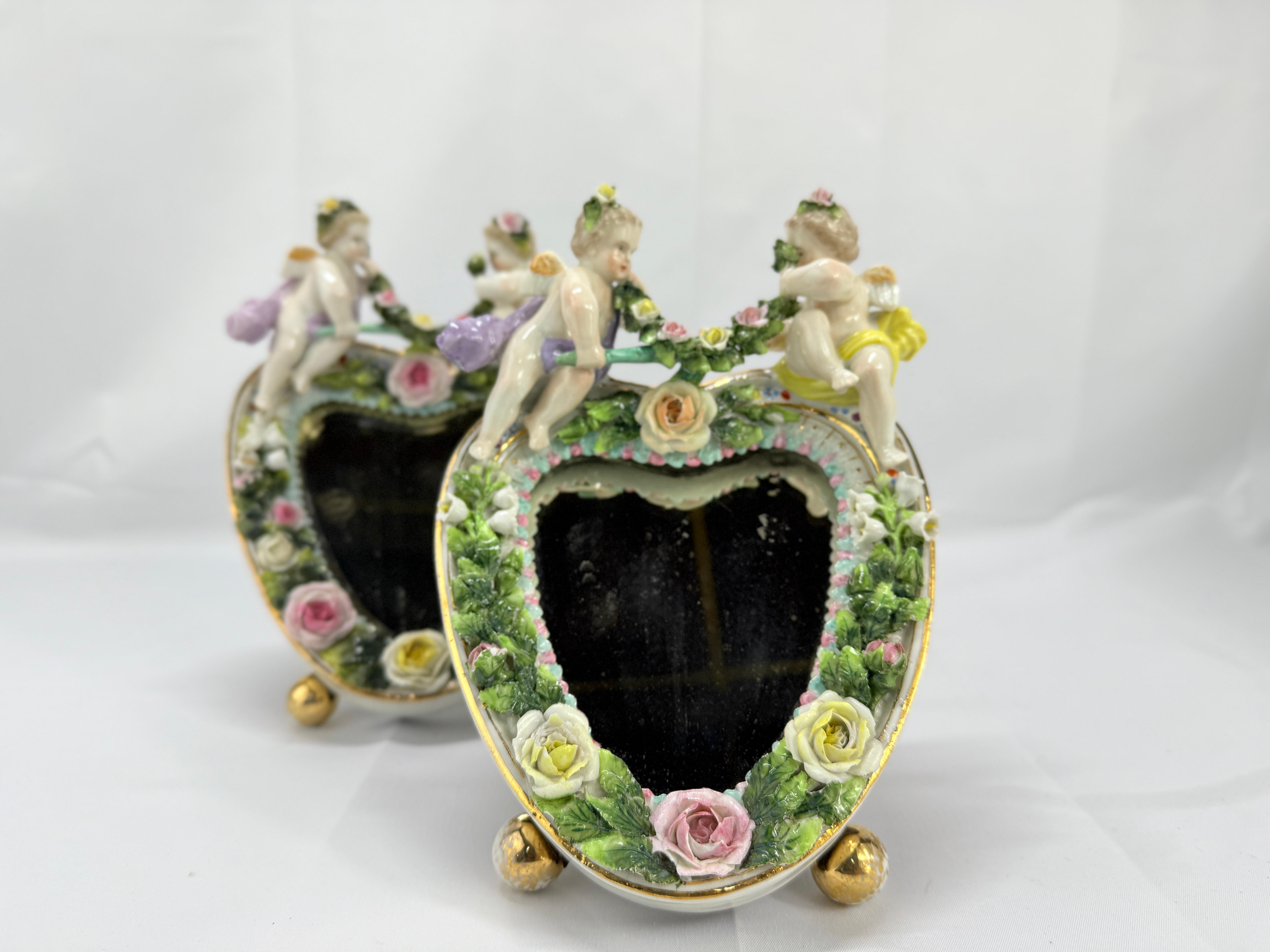 Espejos alemanes de porcelana decorada con pintura, finales del siglo XIX, a la manera de Meissen o Dresden, la cresta con querubines alados sobre flores y hojas aplicadas, la base centrada con un lazo y con dos pies redondos, todo alrededor de una