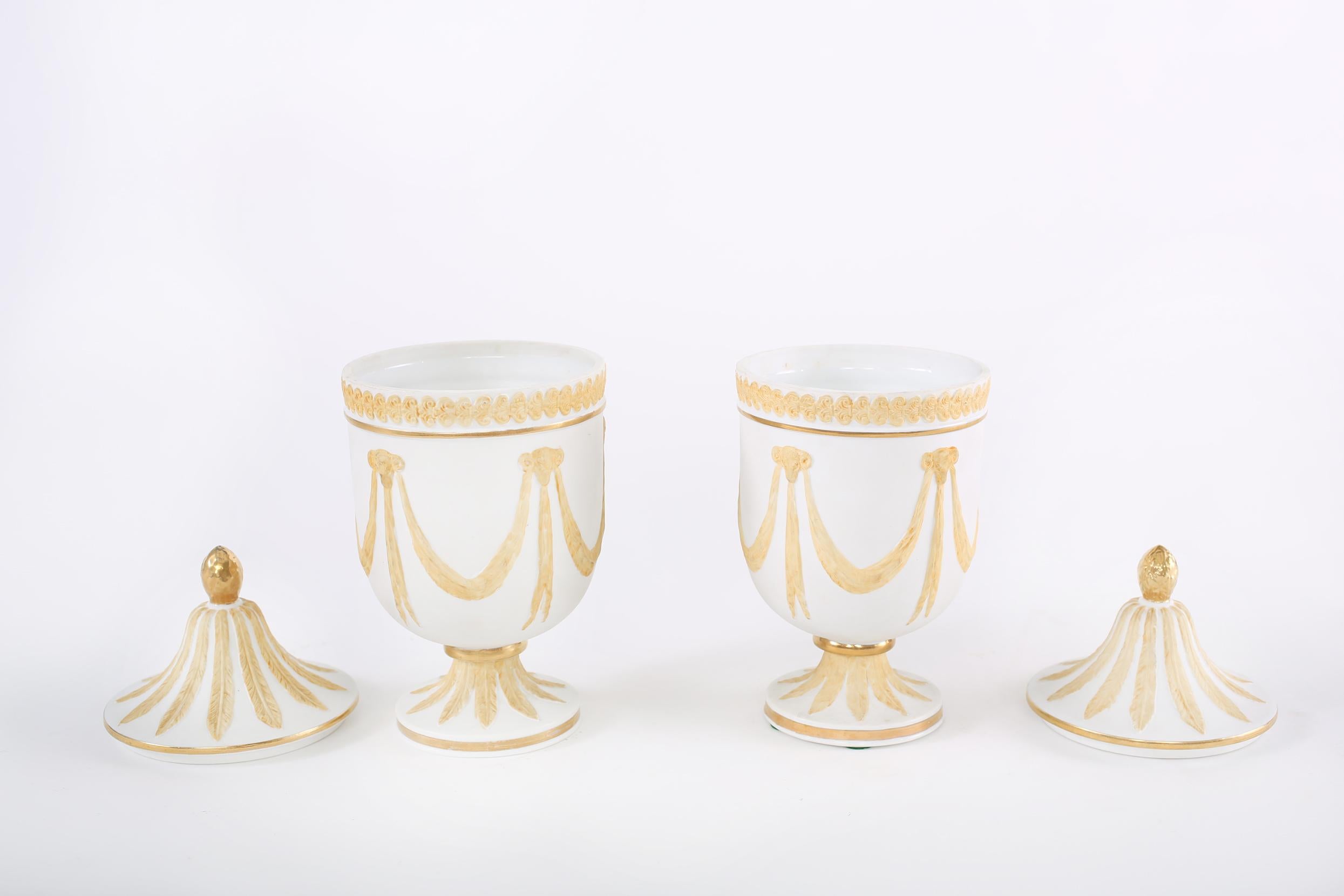 Pair of gilt porcelain decorative covered urns with exterior design details. Each urn is in great condition with minor wear consistent with age / use. Each stands about 10 inches tall x 5 inches diameter.