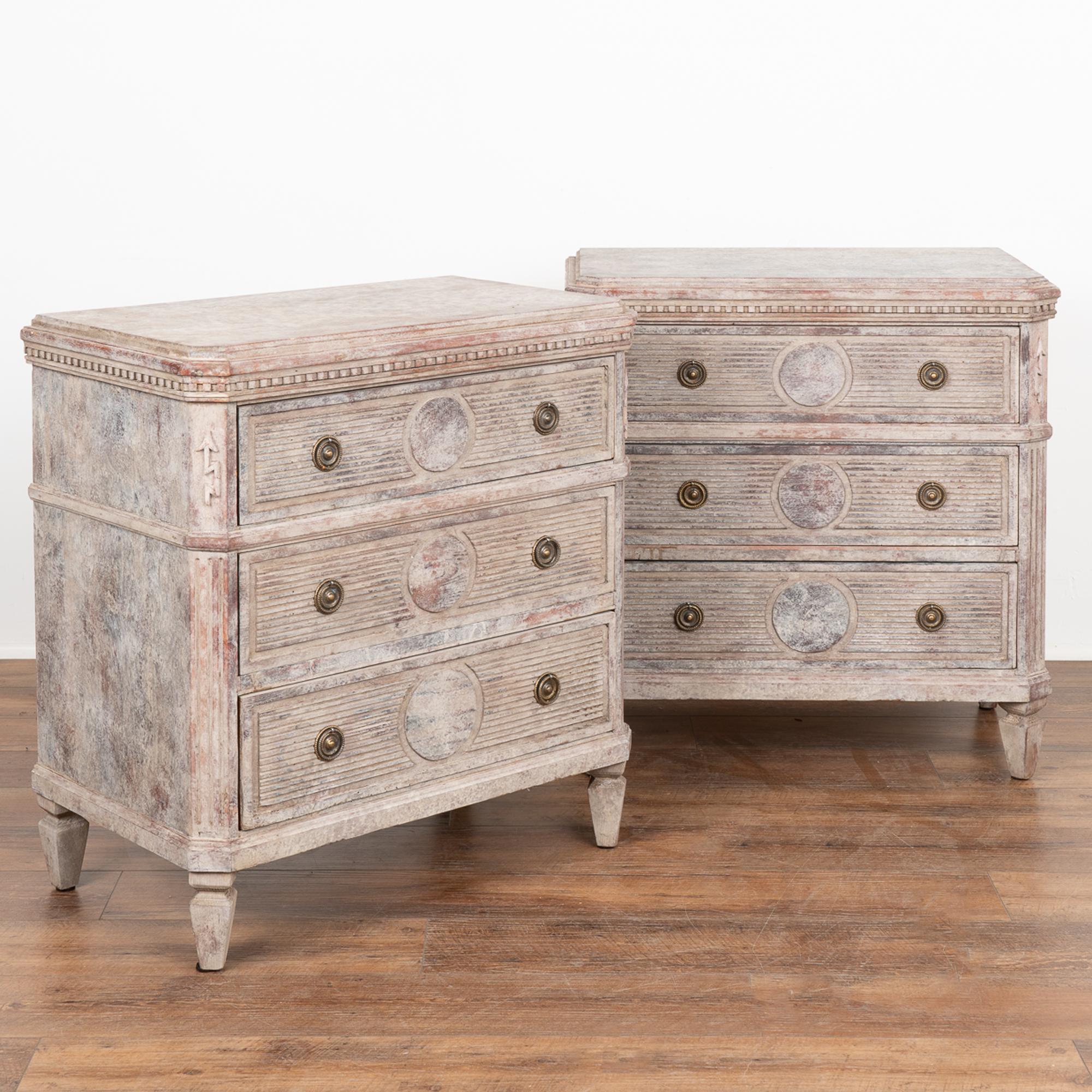 A pair of decorative Gustavian pine chest of drawers painted in mottled shades of  gray and plum.
Canted fluted side posts with unique arrow carving at top, dentil molding, and fluted drawers with circular panel in middle all raised on four tapered