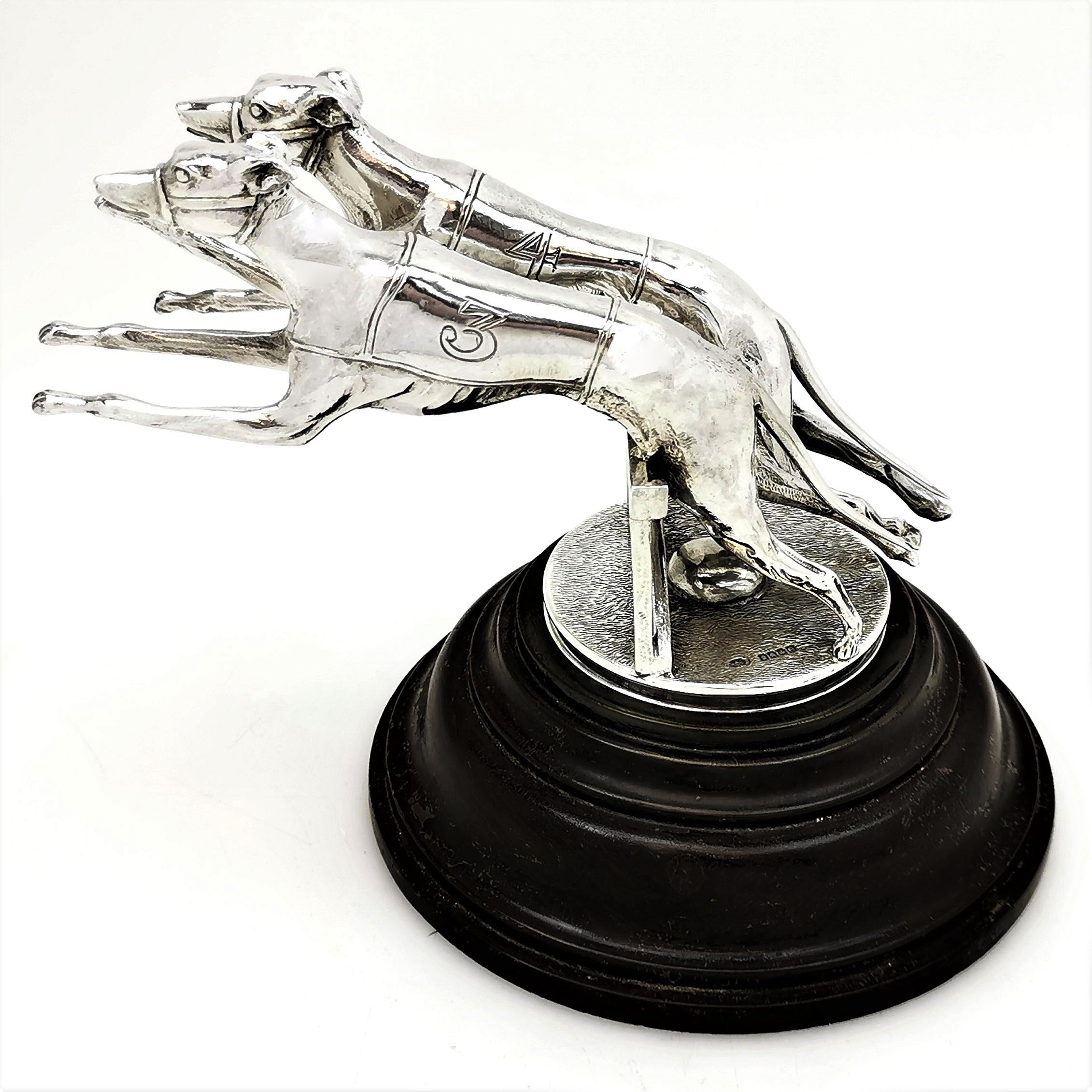 A lovely pair of solid Silver Greyhounds leaping over a hurdle mid race. This sterling silver model of Greyhound Dogs stands on a dark wooden circular plinth. These Greyhounds are created with a wonderful attention to detail, including racing