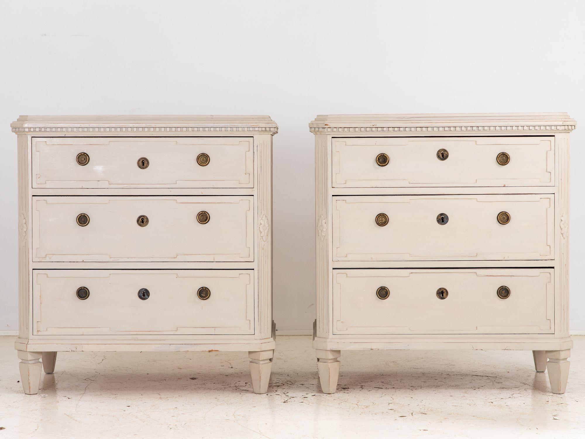 This charming pair of Gustavian-style chests of drawers exudes timeless Swedish elegance. Bathed in a soothing greige paint, they boast three spacious drawers adorned with intricately carved lozenge fronts, adding a touch of sophistication. The
