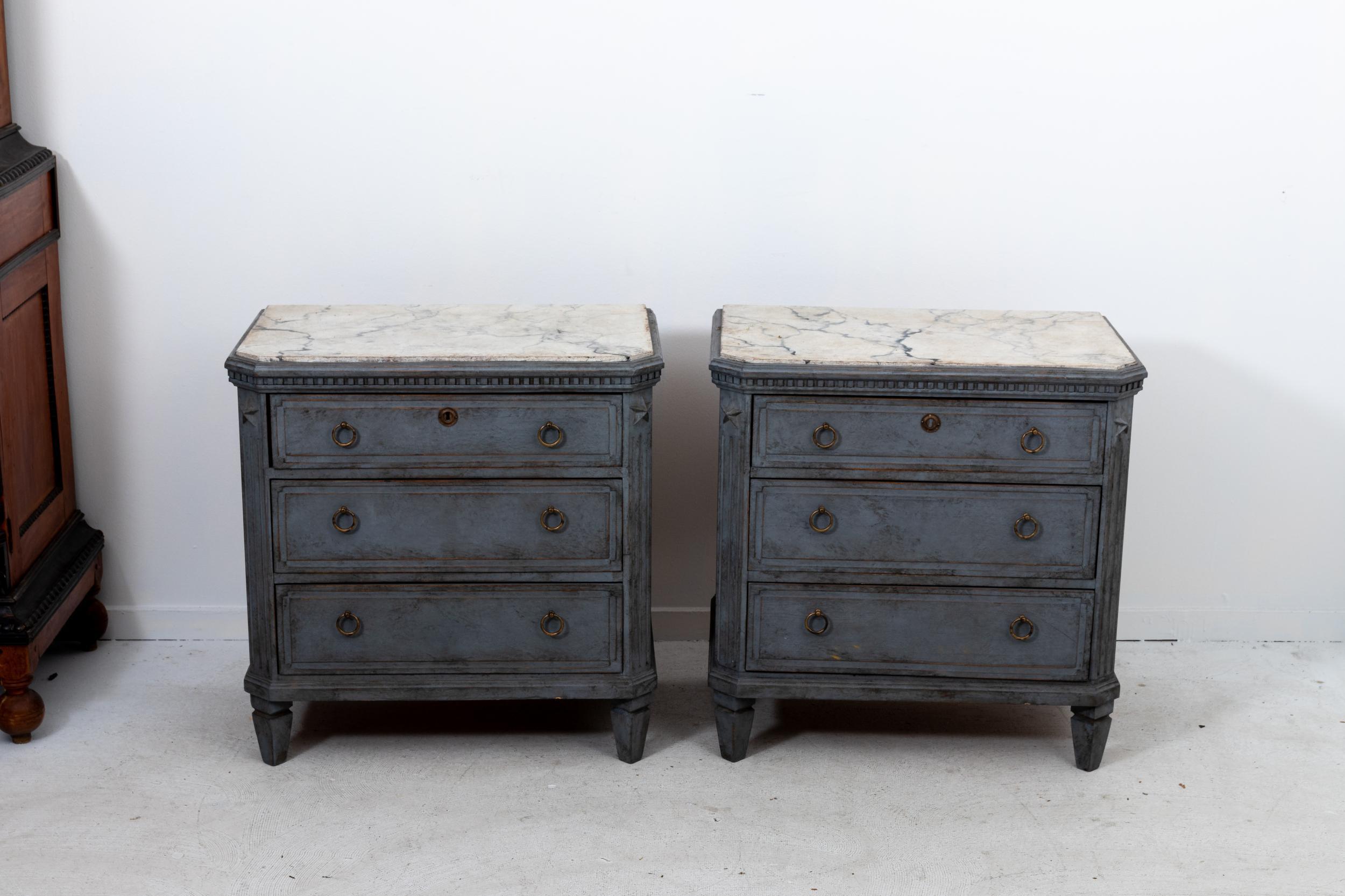 A pair of Gustavian style chests of drawers. A deep gray paint complements the decorative faux marble top. The canted corners are typically of the Gustavian style. Dentil molding, brass hardware, and star medallions complete the look.