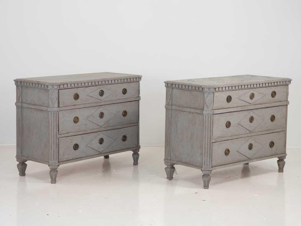 A pair of Neoclassical Gustavian style chests of drawers. Dentil moulding around the top. Canted corners featuring lozenge moulding over fluted legs. Three drawers, each with a center lozenge design. Painted in a classic Swedish Gray.