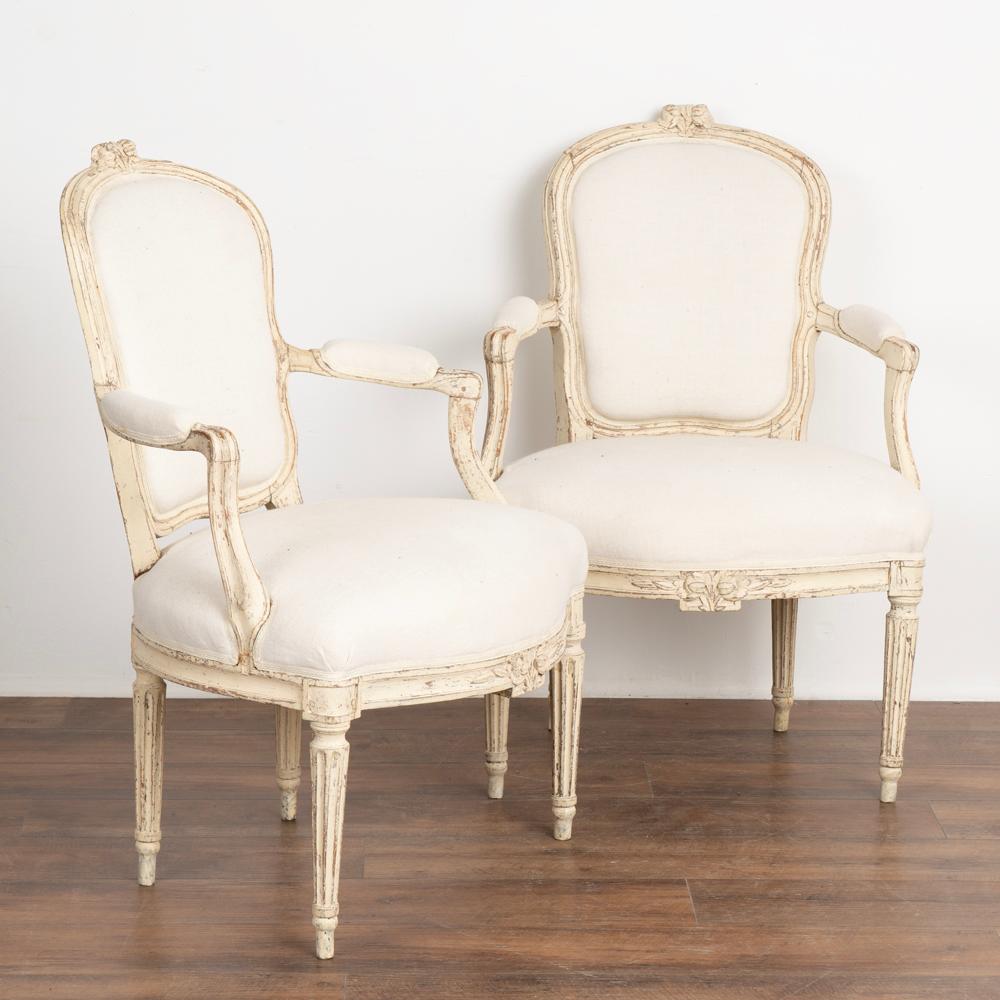 Pair, attractive Swedish country white gustavian painted arm chairs.
Turned, fluted legs and carved accents including floral motif.
White painted finish distressed fitting the age and grace of these lovely arm chairs. 
Restored, stable. Signs of