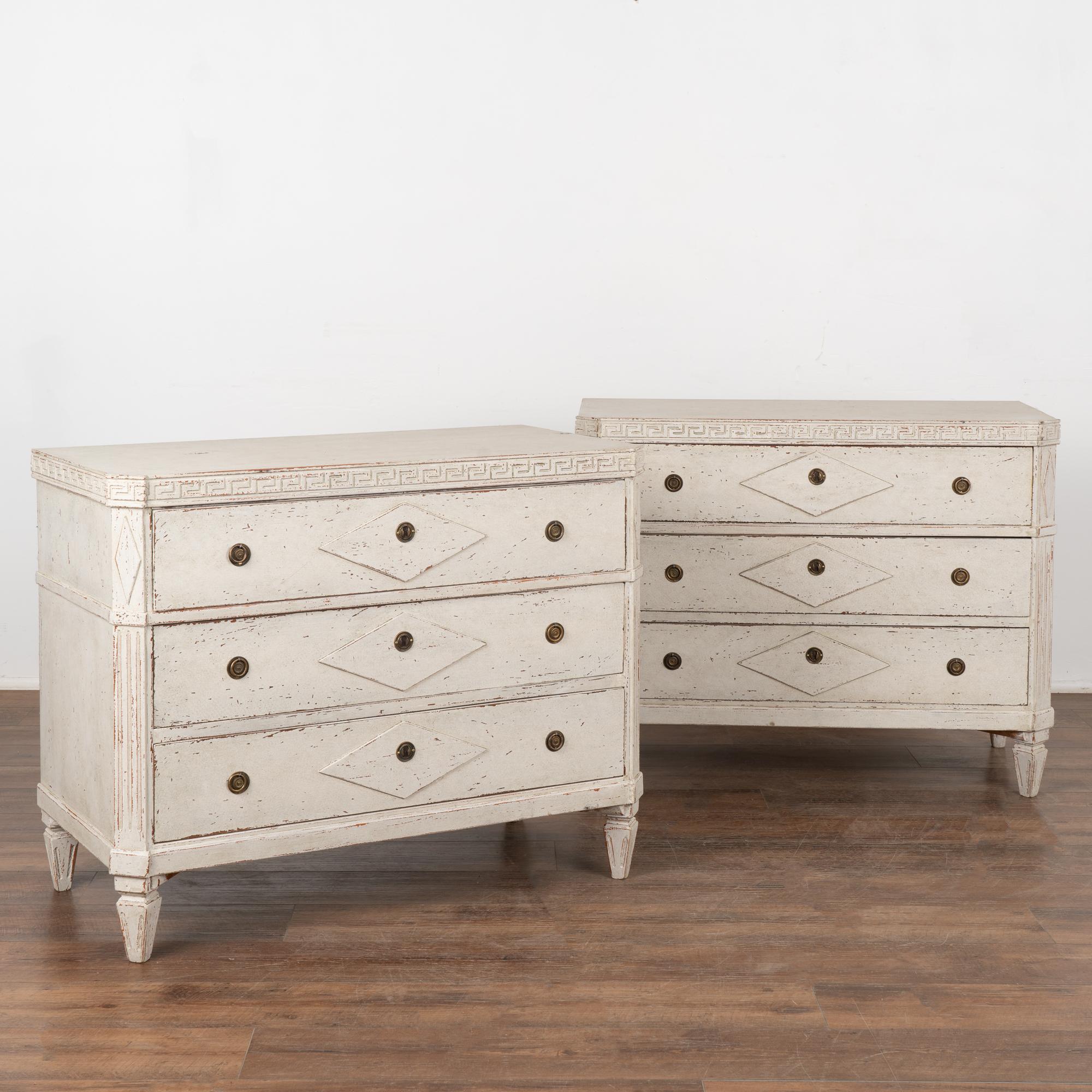 A pair of decorative Gustavian pine chest of drawers painted in shades of  white, fitting their Swedish origin.
Canted fluted side posts with upper carved diamond medallion, raised carved diamond panels along the three drawers, all setting on four