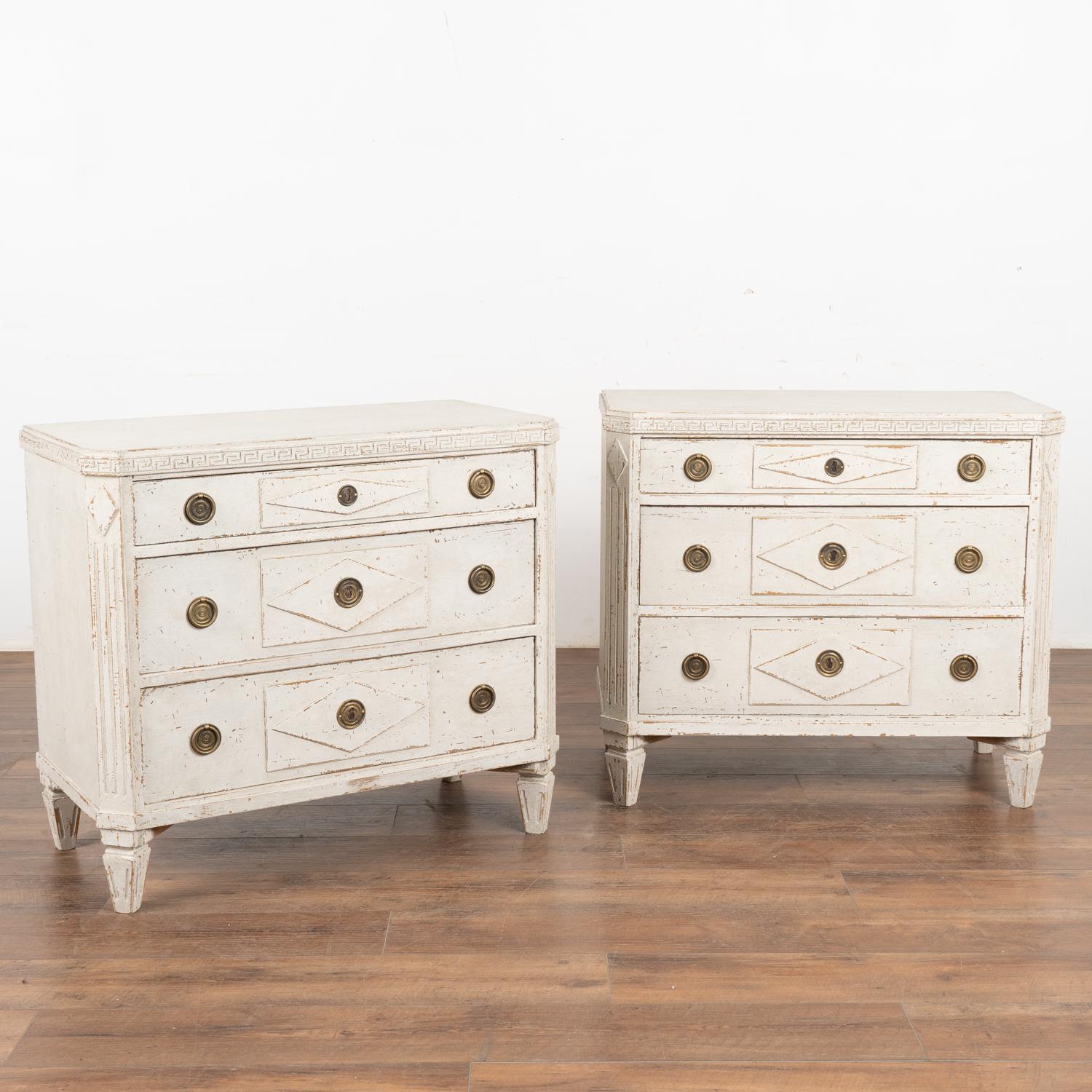 A pair of decorative Gustavian pine chest of drawers painted in shades of  antique white with Greek key motif carved in upper top molding.
Canted fluted side posts with upper carved diamond medallion, raised panels with traditional diamond motif